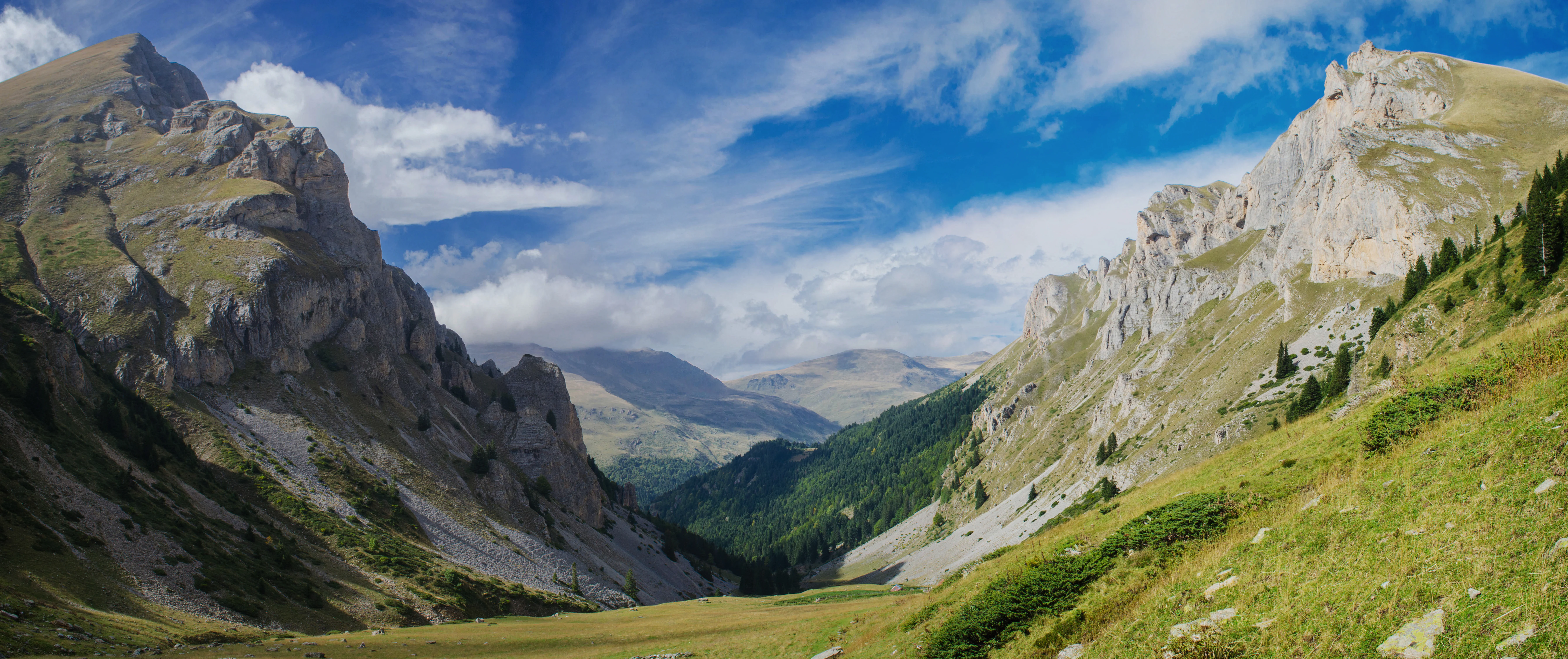 Panoramic view of mountain valley with rocky peaks, green slopes, and blue sky with scattered clouds