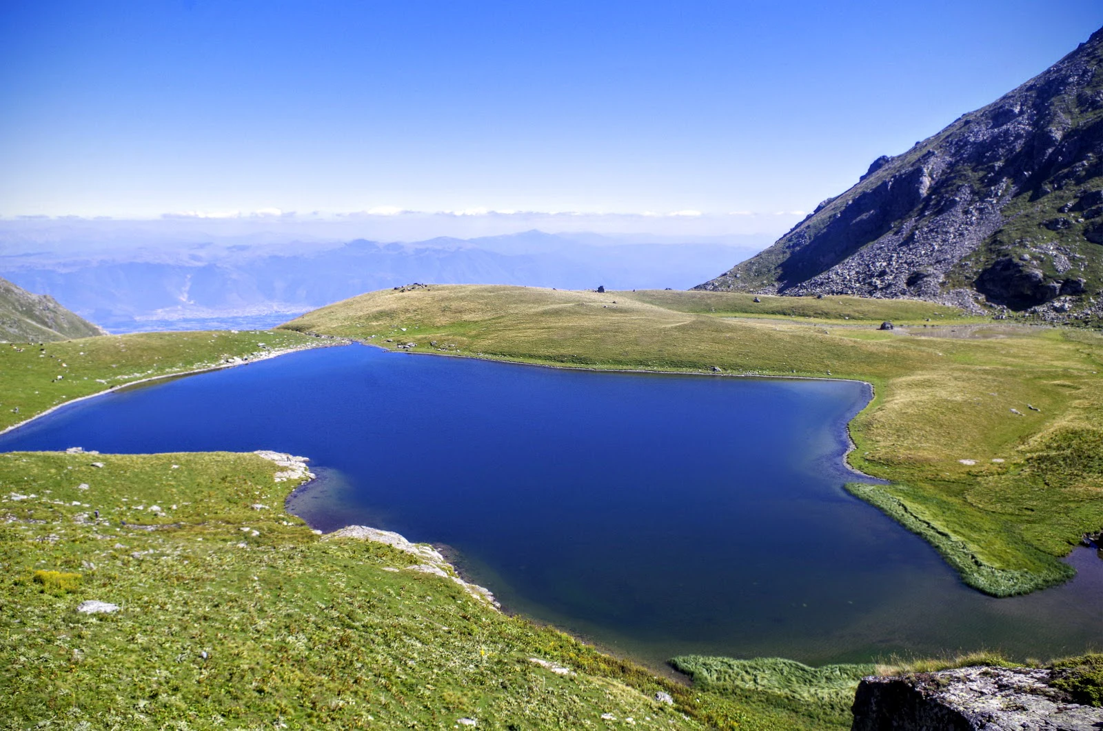 A clear blue glacial lake surrounded by grassy slopes and rocky mountains under a clear blue sky