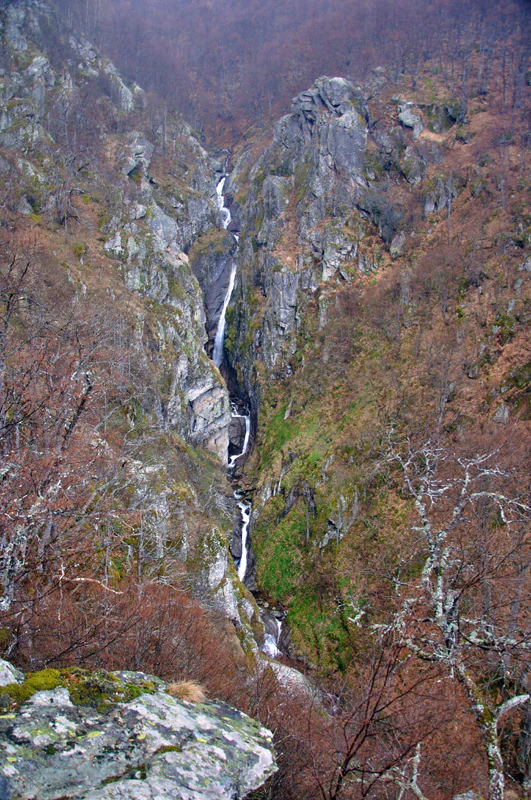 A waterfall flows through a rocky mountain valley with sparse trees and autumn-colored vegetation.