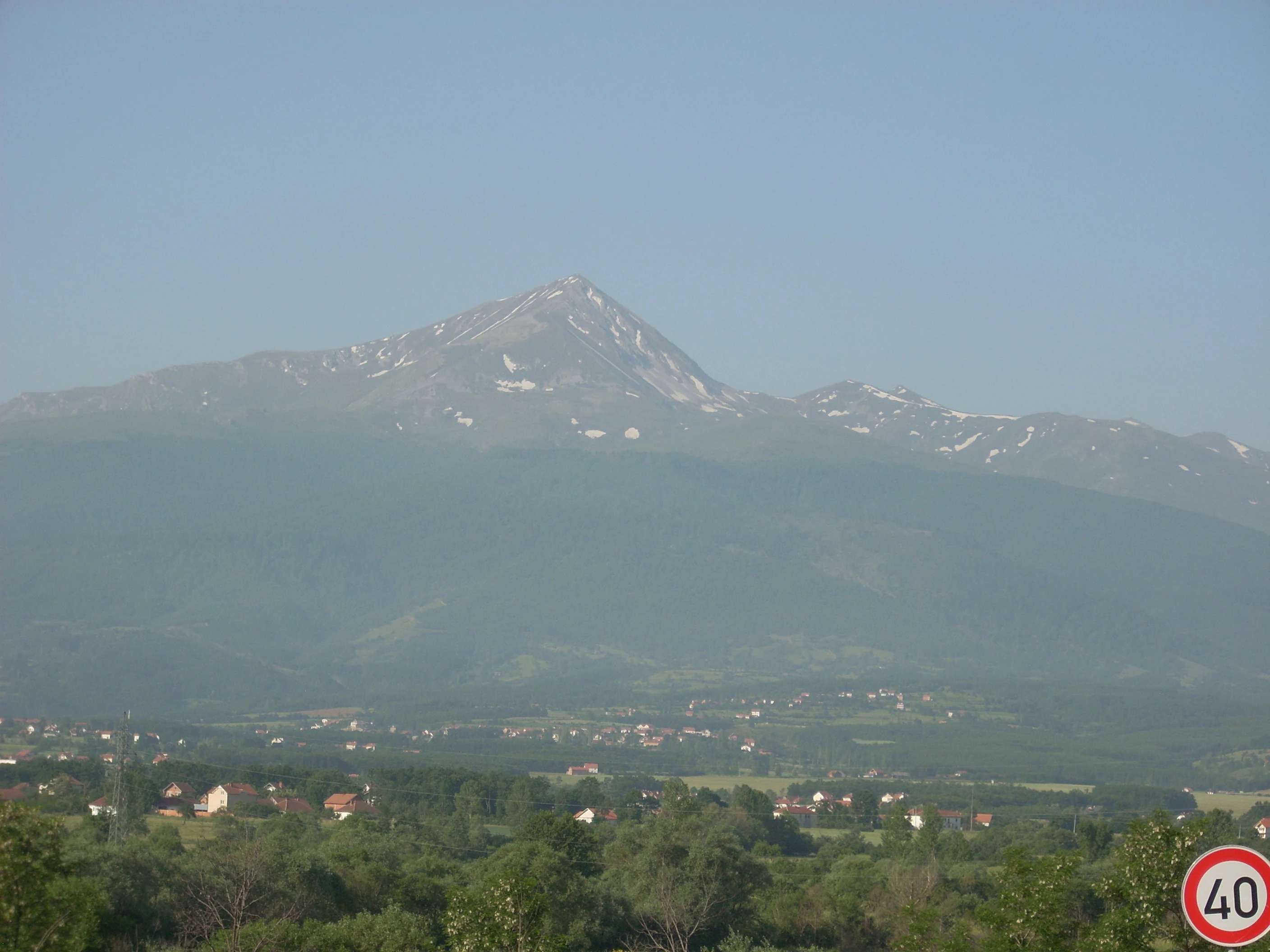 Mountain landscape with prominent peak, green valley below, and red speed limit sign in foreground