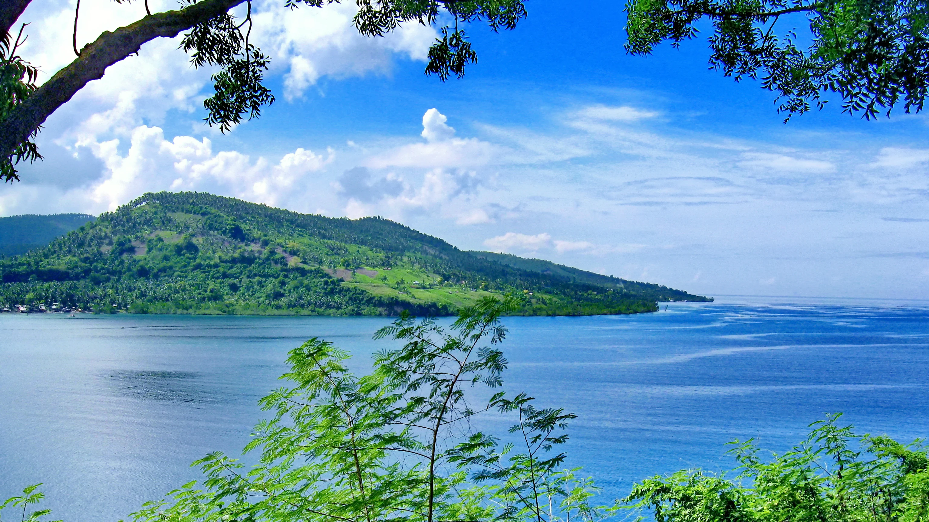 A wide view of a blue bay framed by green trees in the foreground, with a large, lush green mountain peninsula extending into the water under a partly cloudy sky.