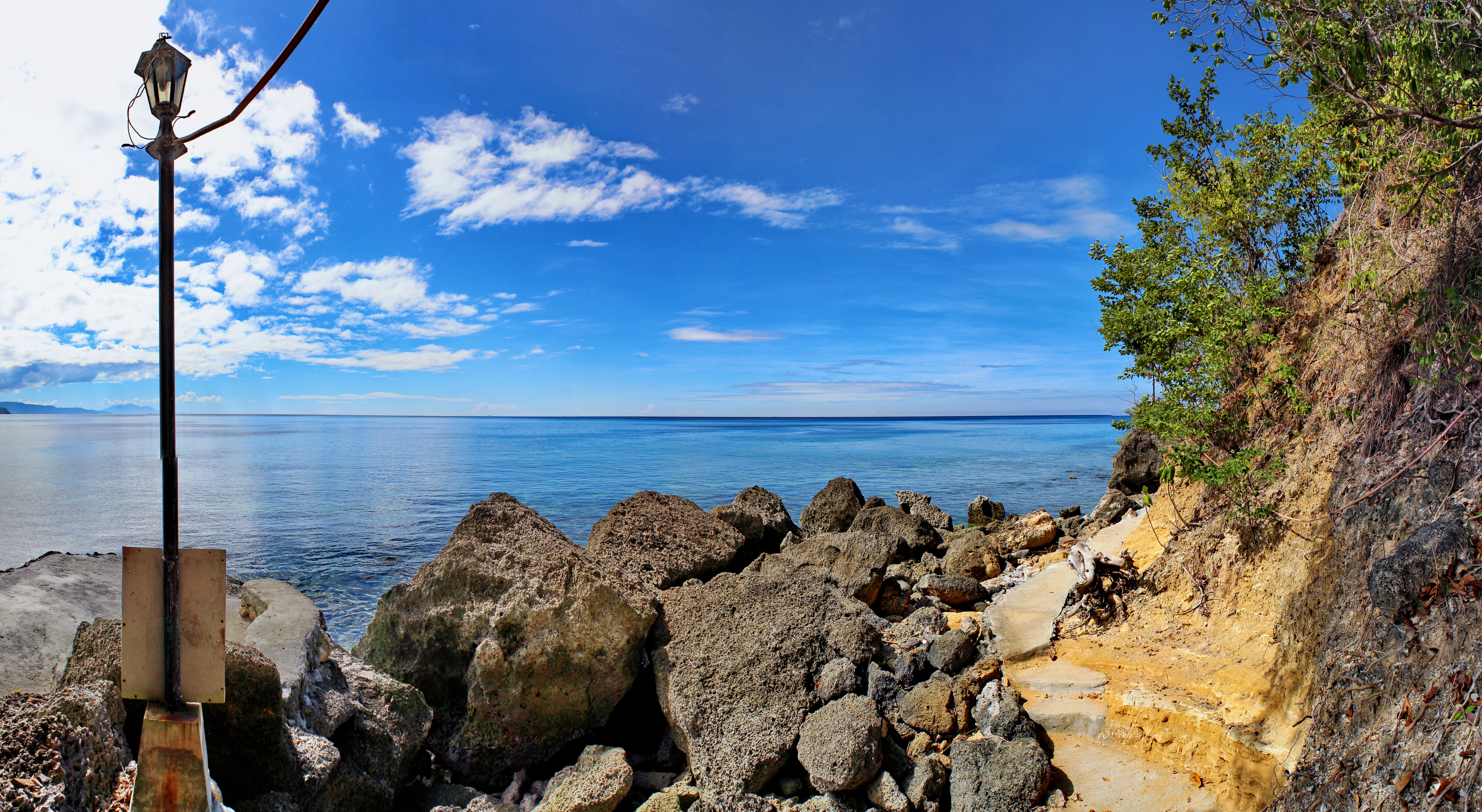 A panoramic view of Sarangani Bay featuring a rocky shoreline, a path running along the edge of a cliff, and a lone lamp post in the foreground under a bright blue sky.