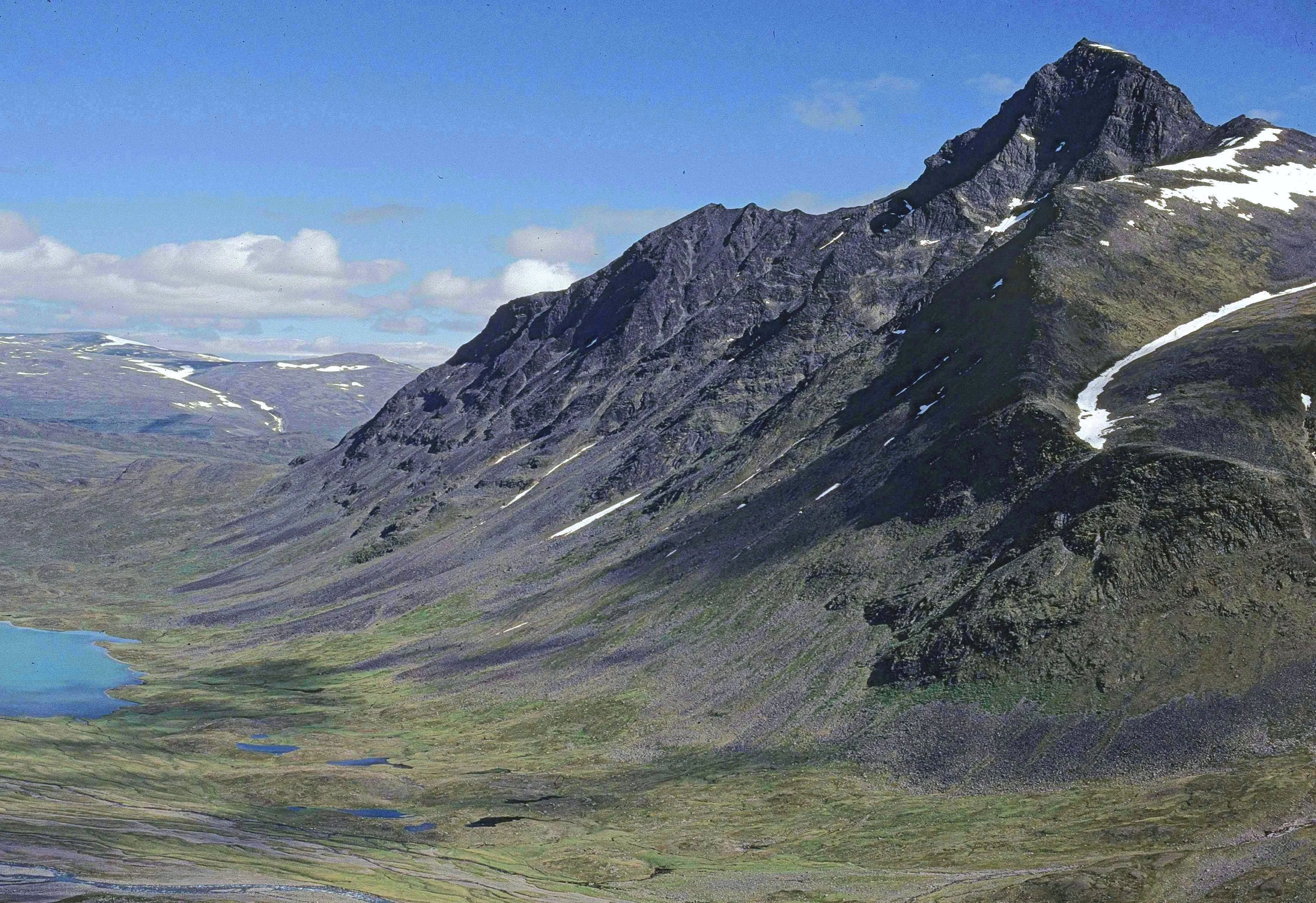 Wide view of a mountain landscape with a blue lake in the valley, snow patches on rocky peaks, and clear sky