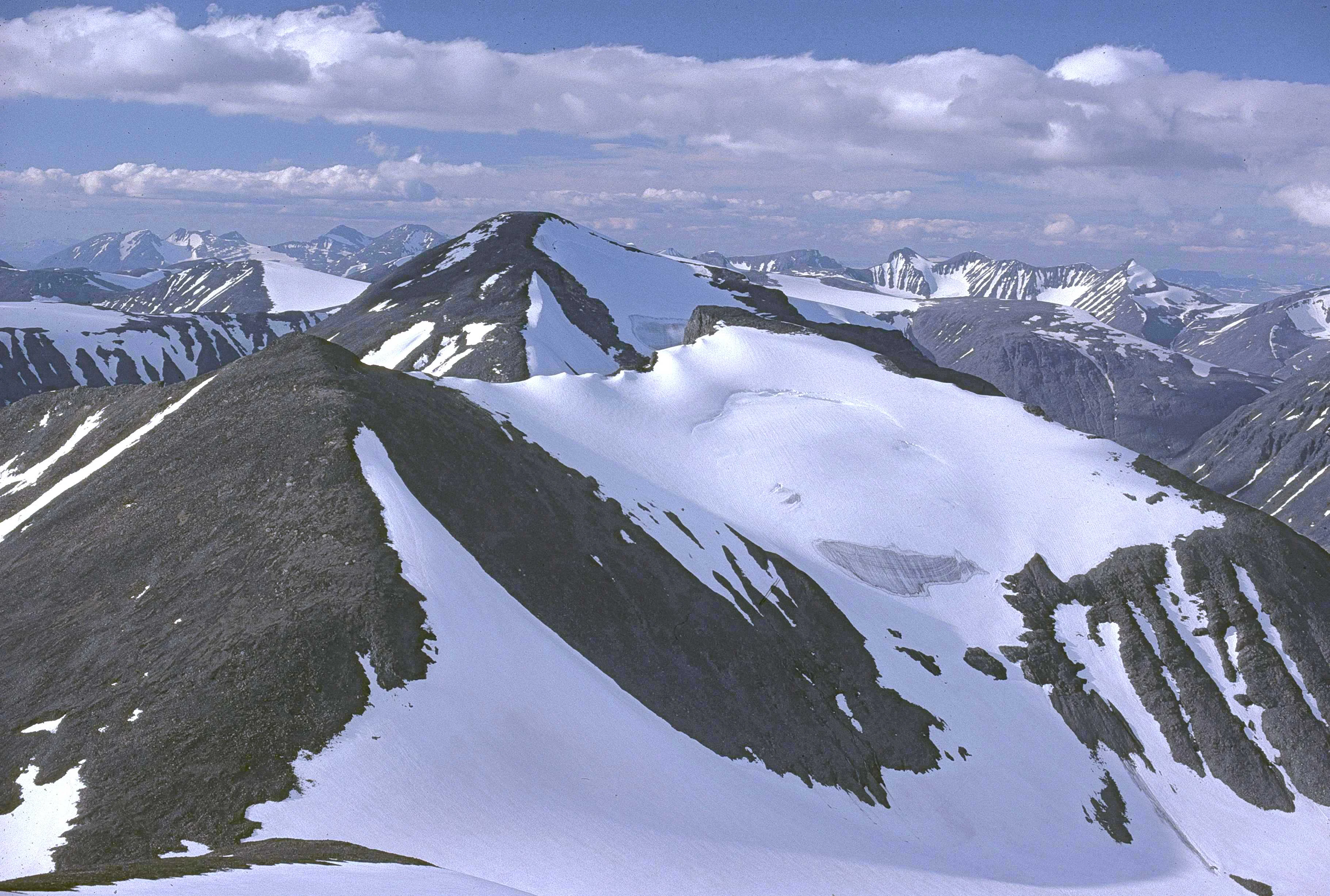 Snow-covered mountain peaks with patches of green vegetation, distant mountain ranges under a partly cloudy sky