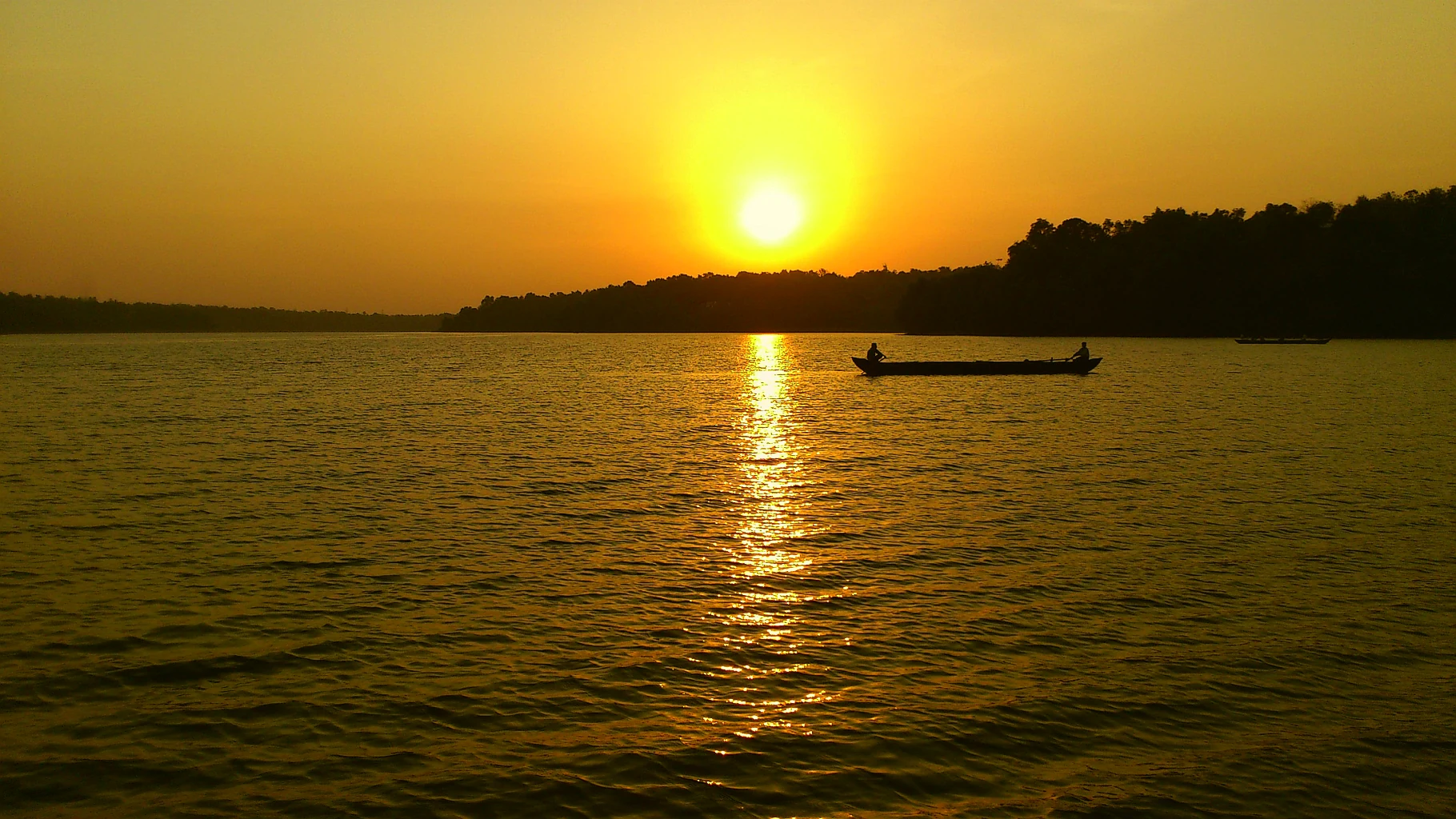A wide view of a calm, large lake at sunset with the sun low on the horizon, casting a golden glow on the water and a silhouette of a small boat.
