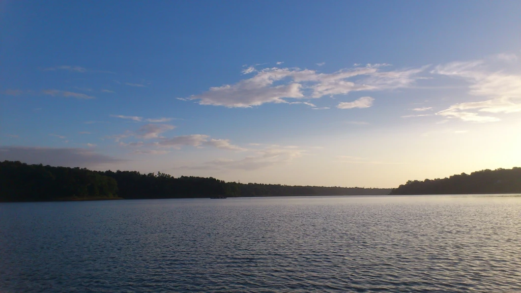 A wide view of a calm, large lake under a clear blue sky with soft sunset light hitting the horizon, surrounded by dark treelines on both shores.