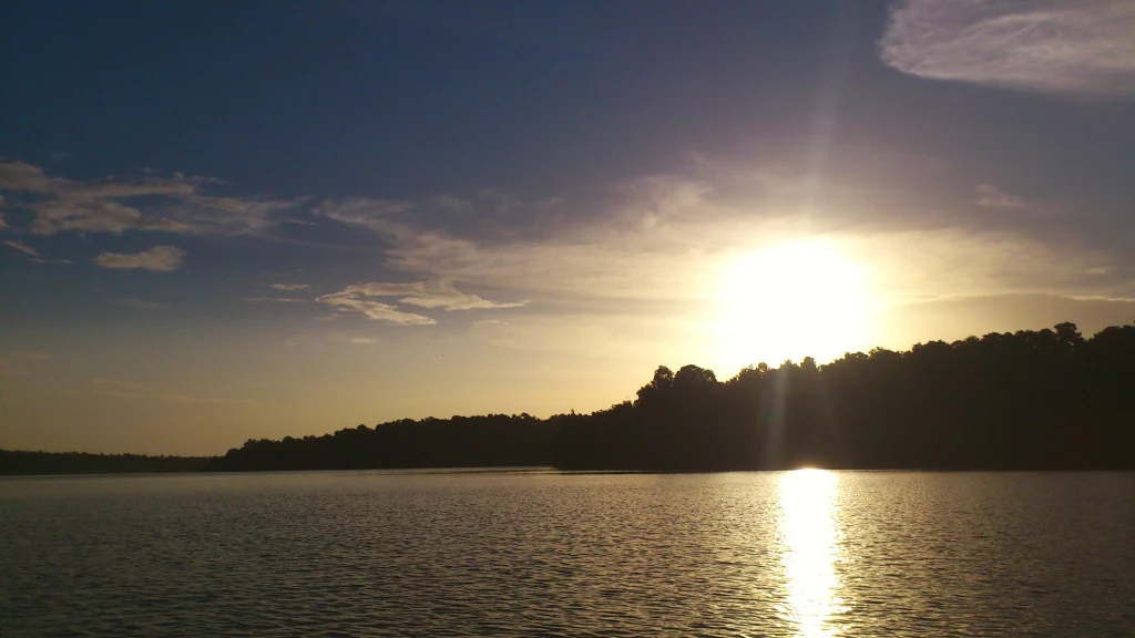 A wide angle sunset view over a calm lake, with the silhouette of a forested shoreline and the sun low in the sky.