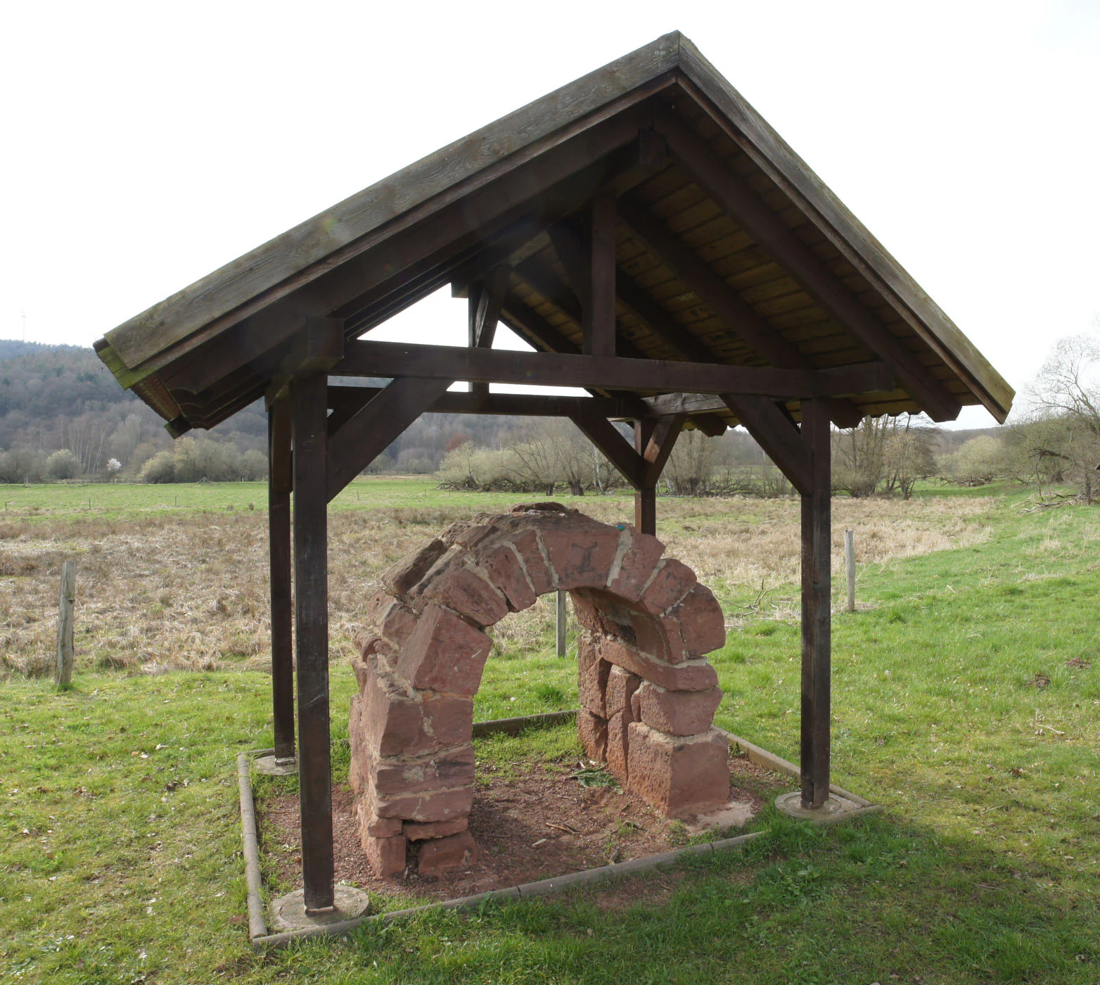 A small, open-sided wooden pavilion protecting a reconstructed sandstone arch structure located in a grassy field.