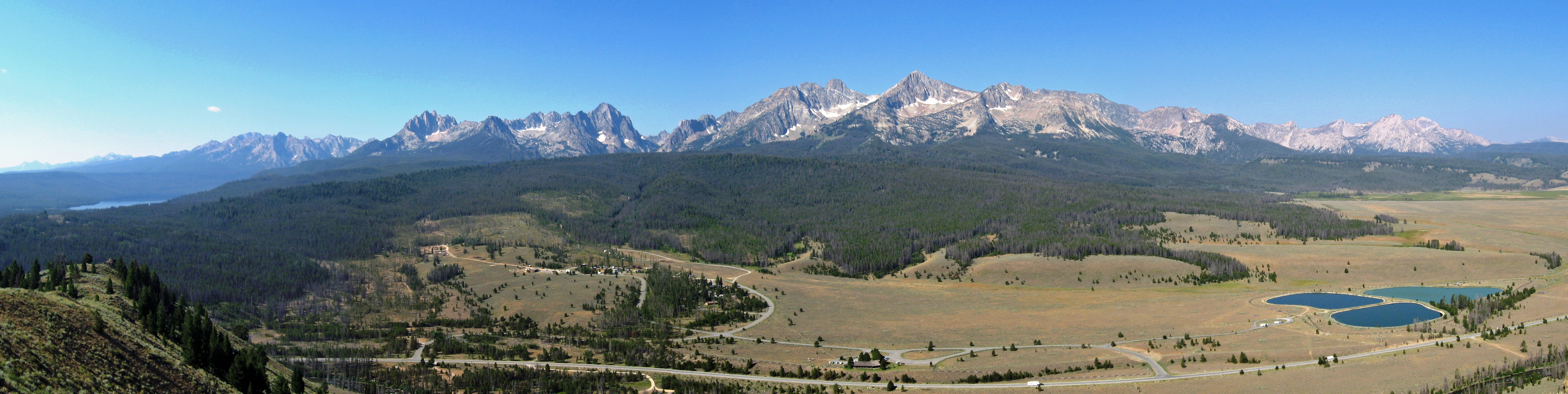 A wide panoramic landscape view featuring a jagged mountain range in the distance, a large pine forest at the base of the mountains, and a flat, open valley in the foreground with small ponds.