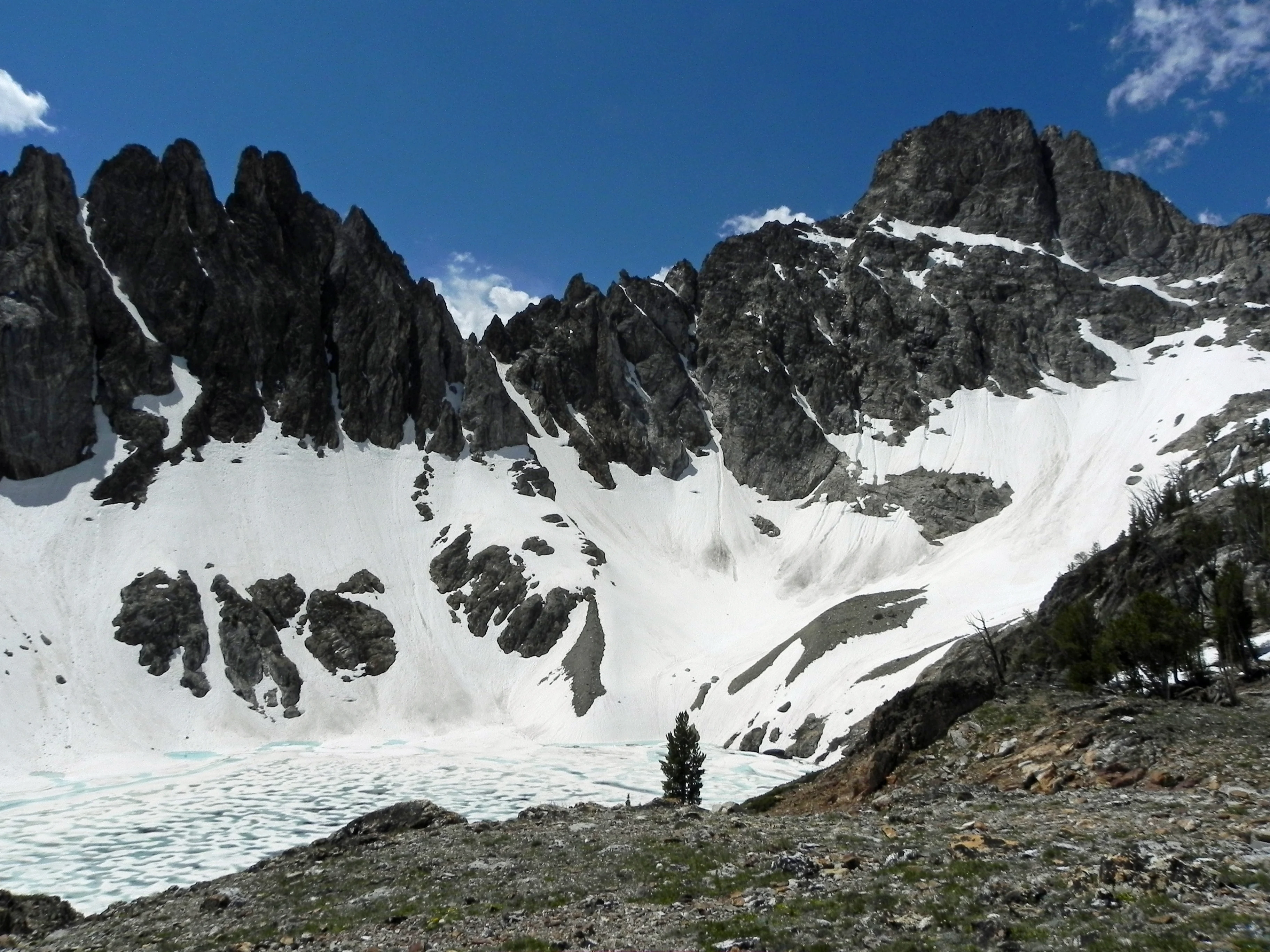 A jagged, dark granite mountain peak rises above a steep basin filled with patches of white snow and a partially frozen alpine lake.