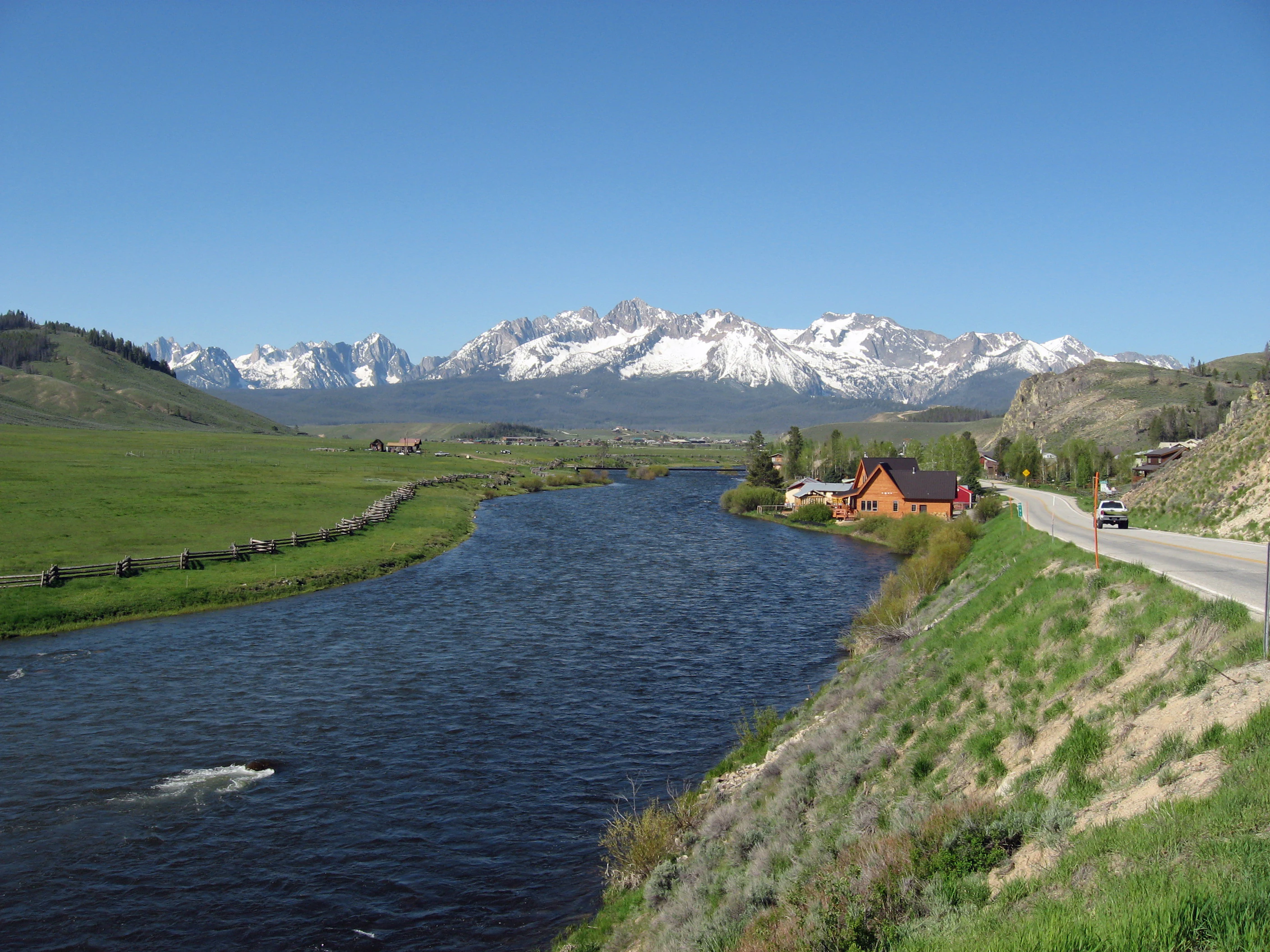 A wide view of the Sawtooth Mountain Range with snow-capped peaks in the background, overlooking the Salmon River flowing through a grassy valley with a small house and a road in the foreground.