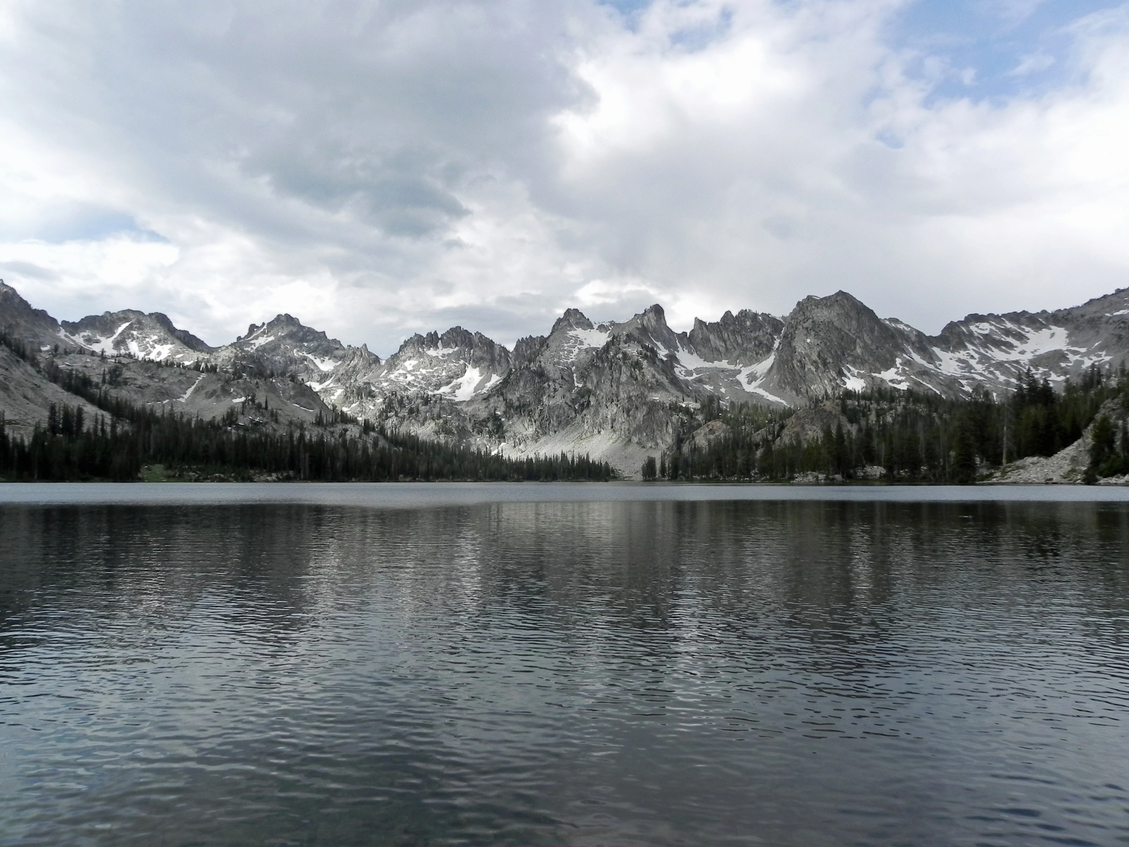 A wide view of a calm, dark lake reflecting snow-dusted, rugged mountain peaks under a cloudy sky.