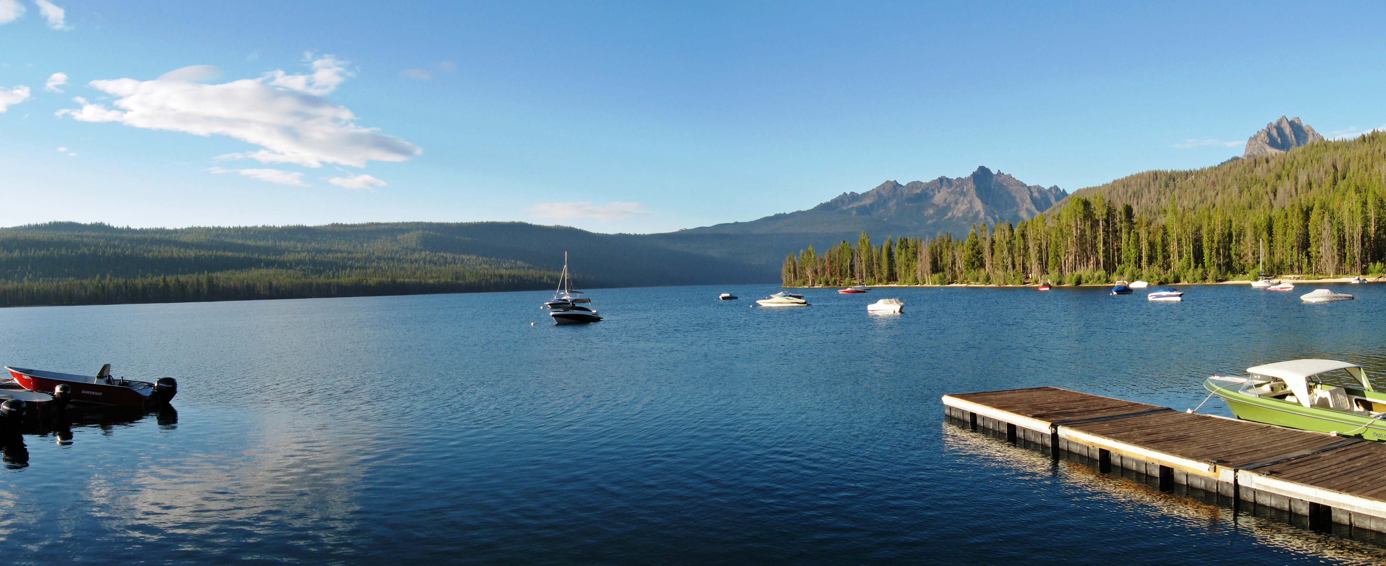 A calm, wide lake with several small boats moored on the water and a wooden dock in the foreground; pine forest and jagged mountain peaks line the distant shore under a clear blue sky.
