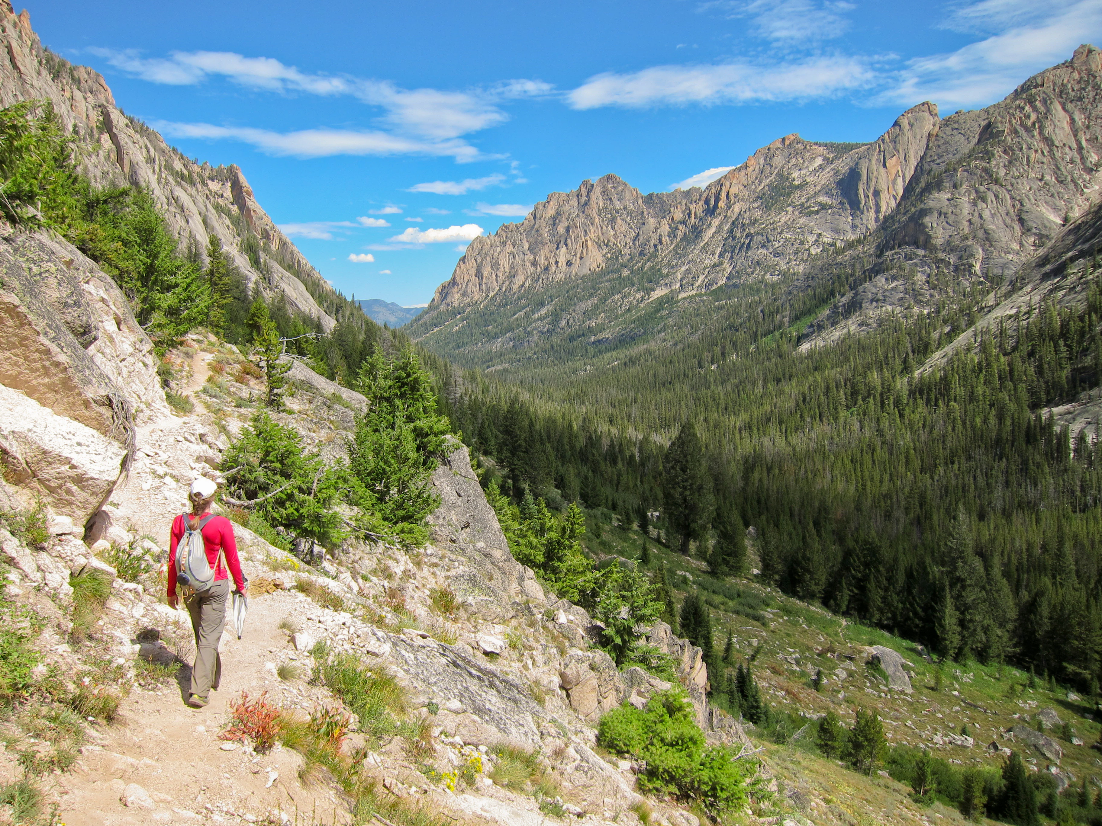 A hiker wearing a red shirt and backpack walks along a rocky mountain trail overlooking a deep, forested canyon flanked by craggy peaks.