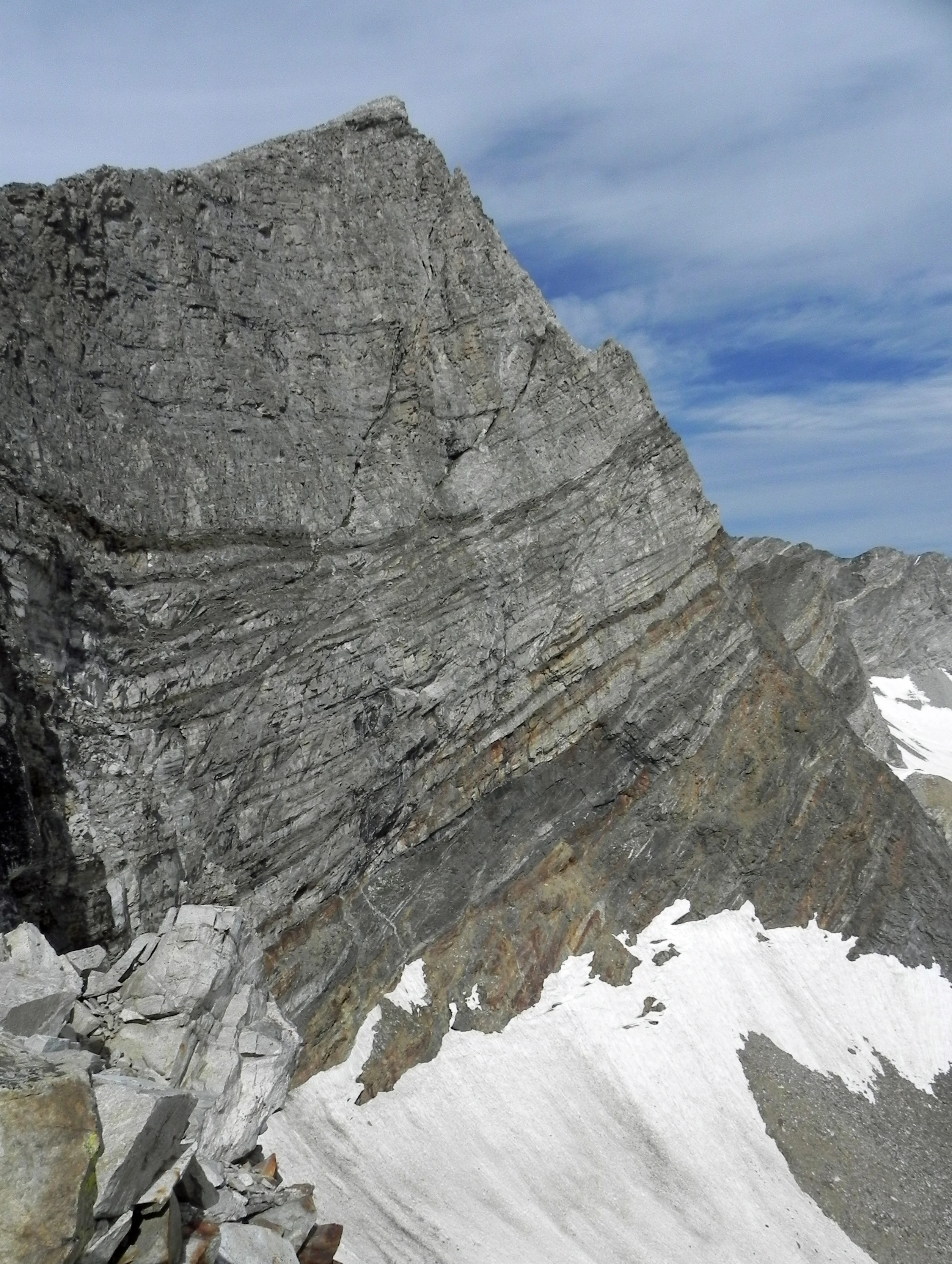 A close-up view of a steep, craggy mountain face showing layered rock strata and patches of snow at the base.