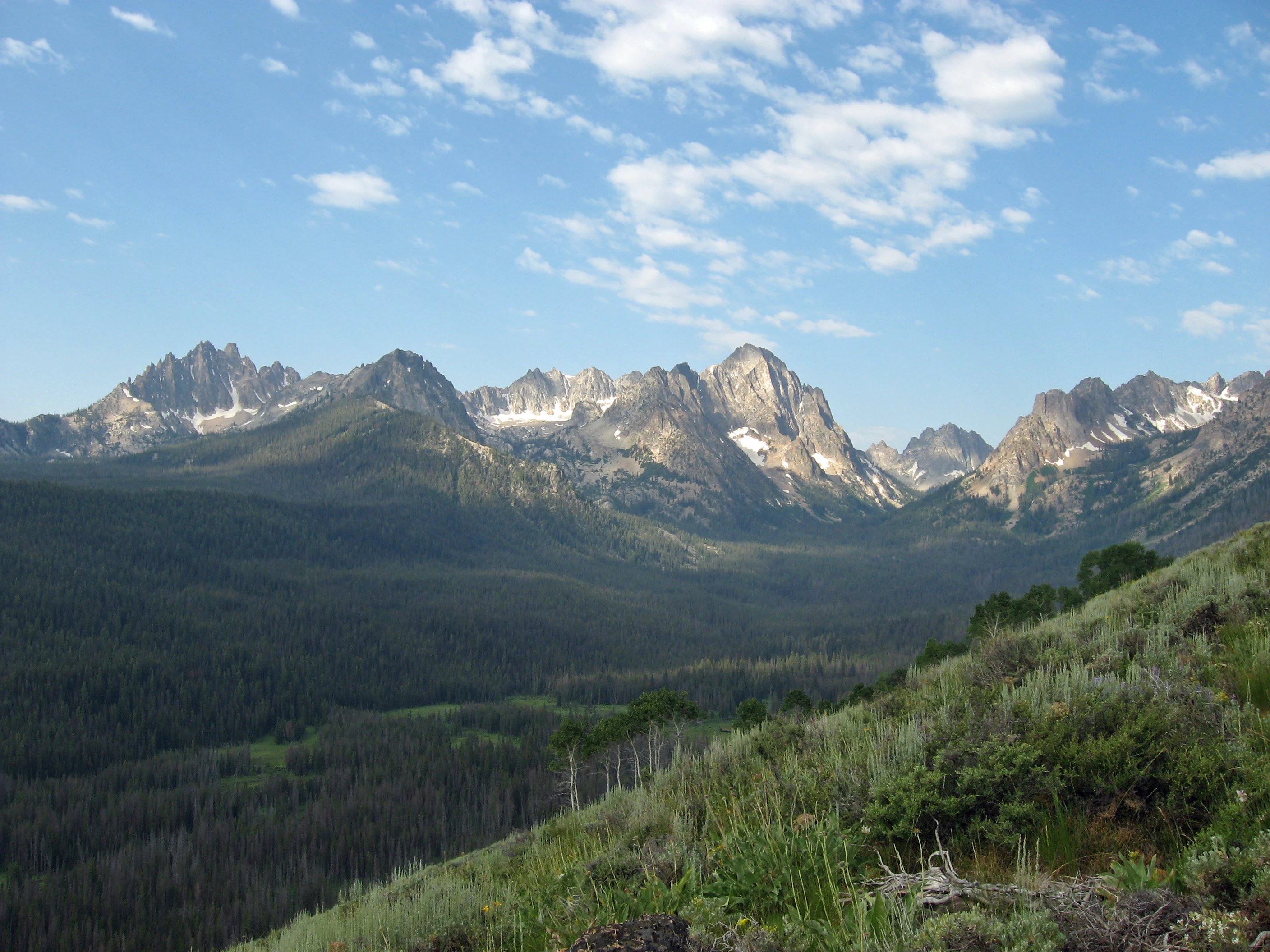 Rugged, jagged mountain peaks with small patches of snow rise above a deep, forested valley under a bright blue sky with scattered clouds.