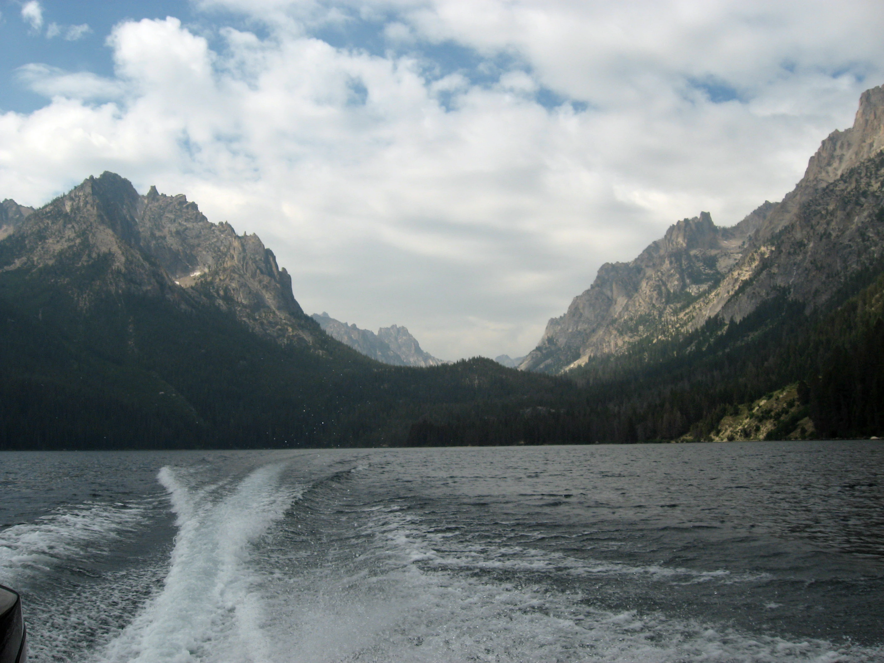 A wide view of a large alpine lake with a prominent boat wake in the foreground, leading toward steep, rocky mountains under a cloudy sky.