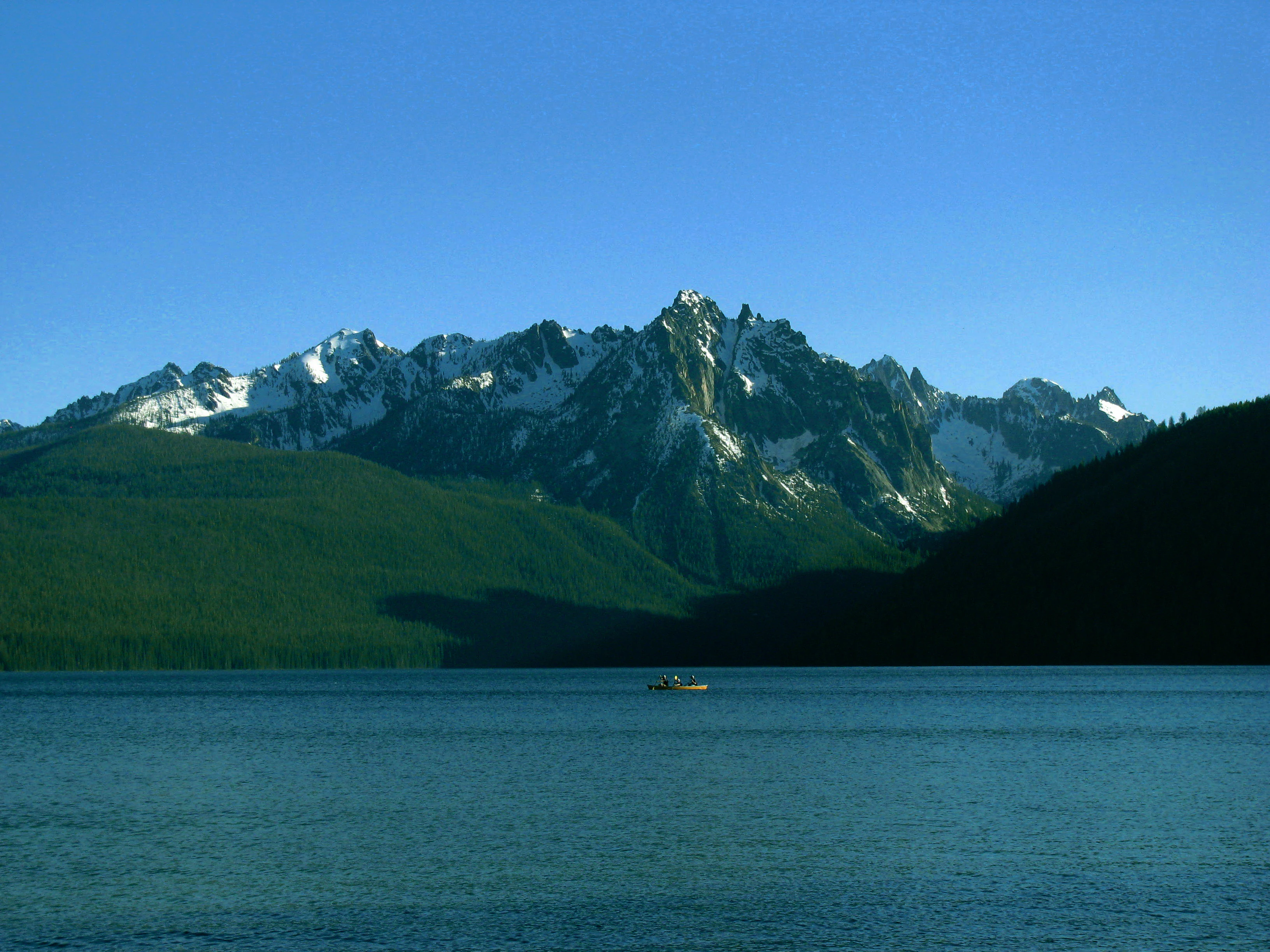 A wide view of Redfish Lake in the foreground, with the snow-capped, jagged peaks of the Sawtooth Range rising in the background under a clear blue sky.