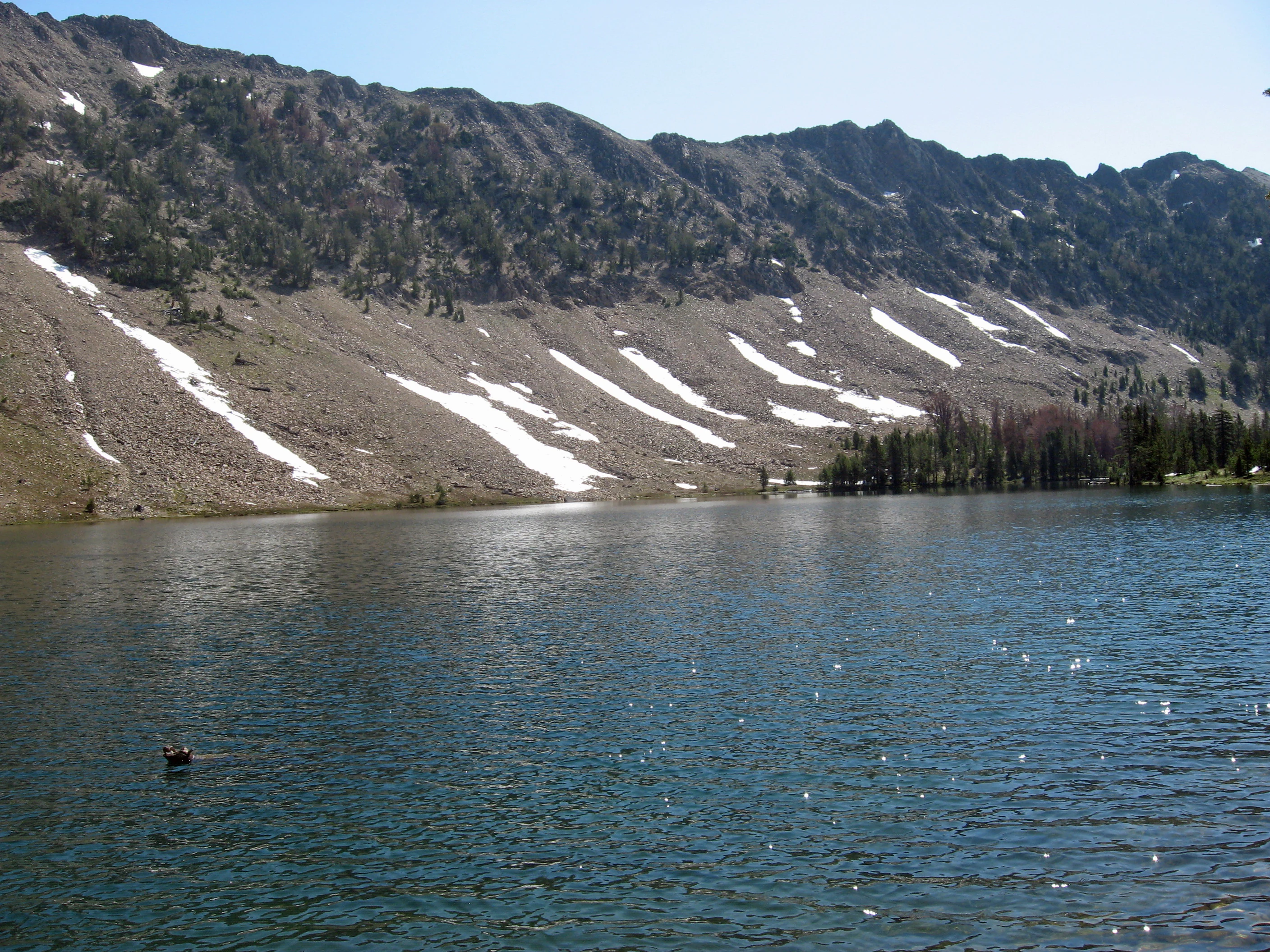 A clear alpine lake reflects a rocky mountain slope with streaks of snow across its face.