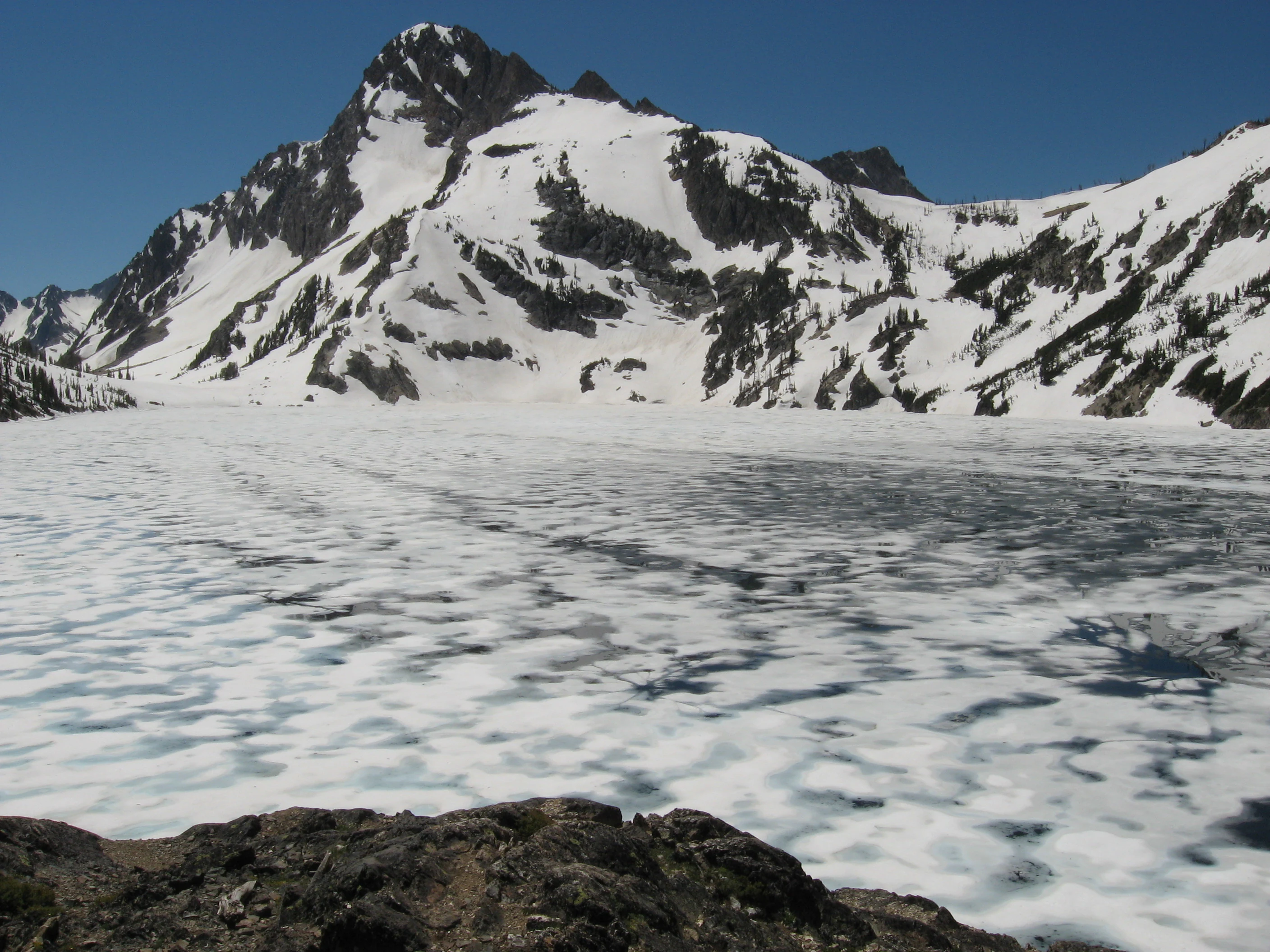 A wide angle shot of a large alpine lake mostly covered in white ice, with a tall, snow-capped mountain rising directly behind it against a clear blue sky.