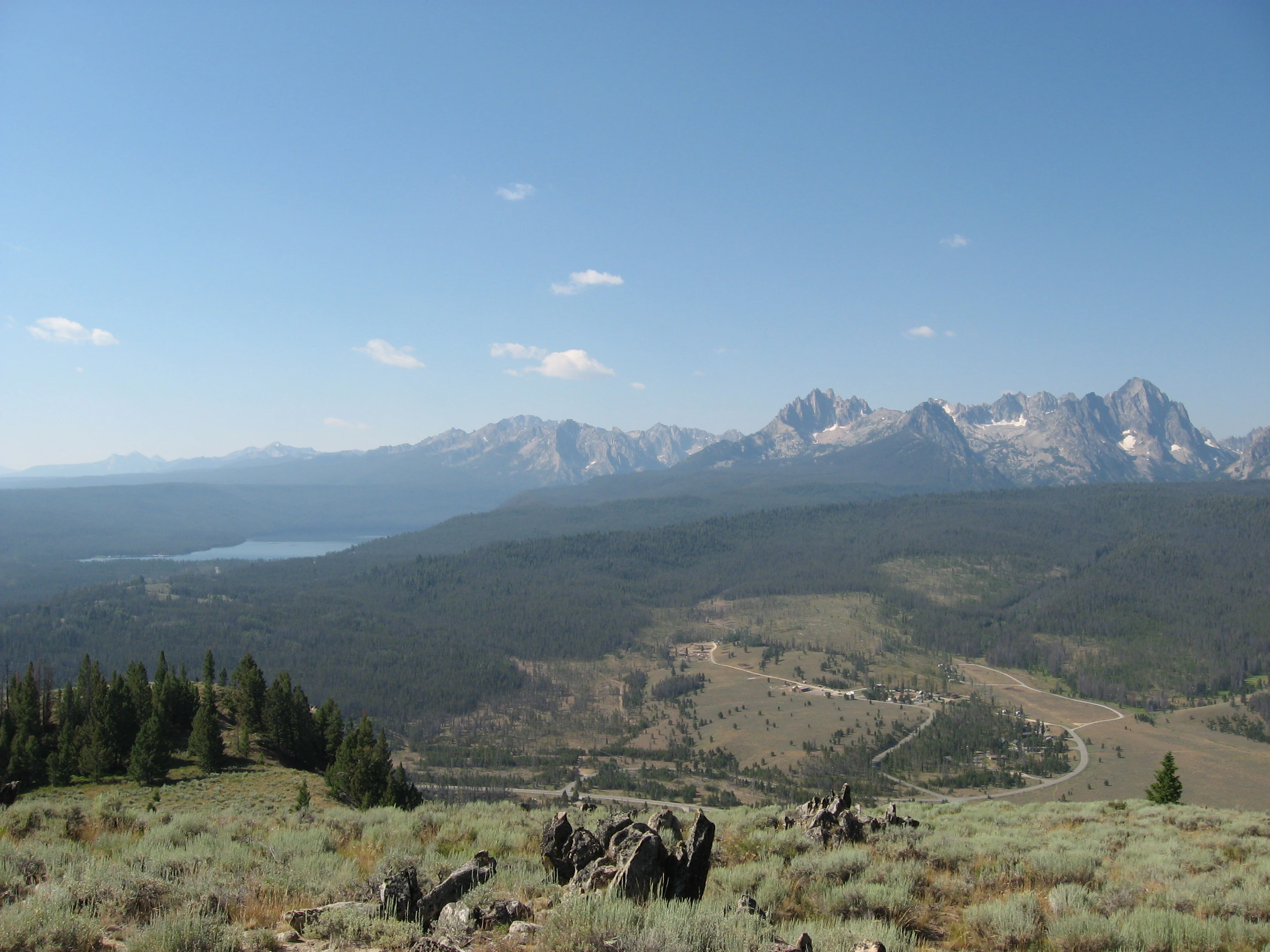 A high angle panoramic view of the rugged Sawtooth mountain range with snow-dusted peaks, overlooking a forested valley with a visible lake in the distance.