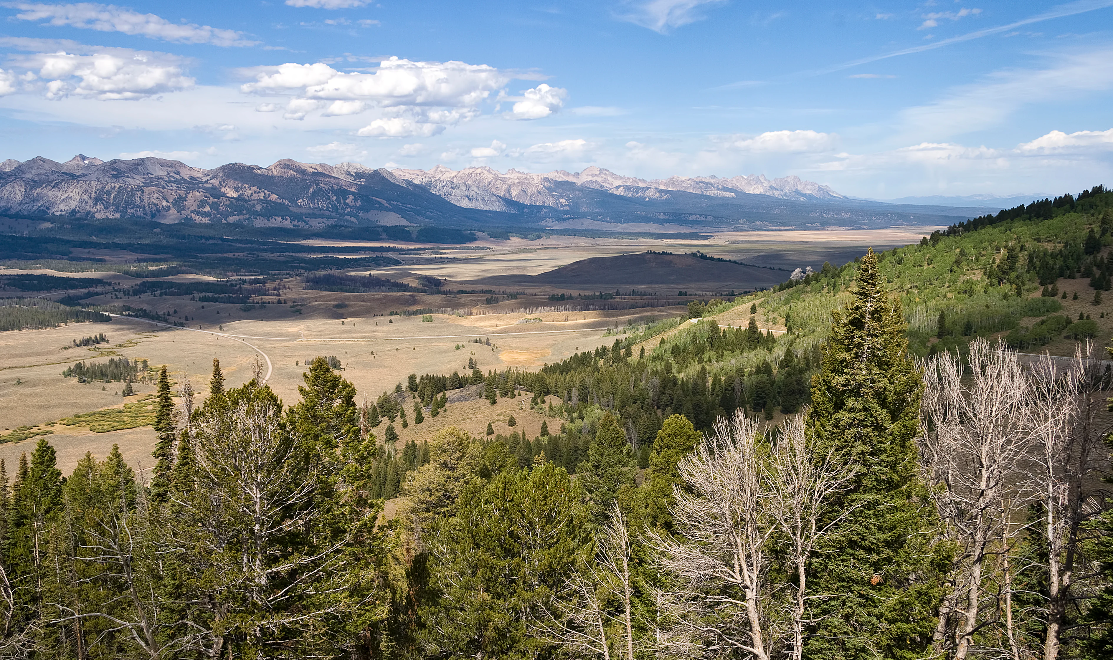 Panoramic landscape view from a high vantage point overlooking a wide valley floor, with rugged mountain ranges in the distance under a blue sky with scattered clouds.