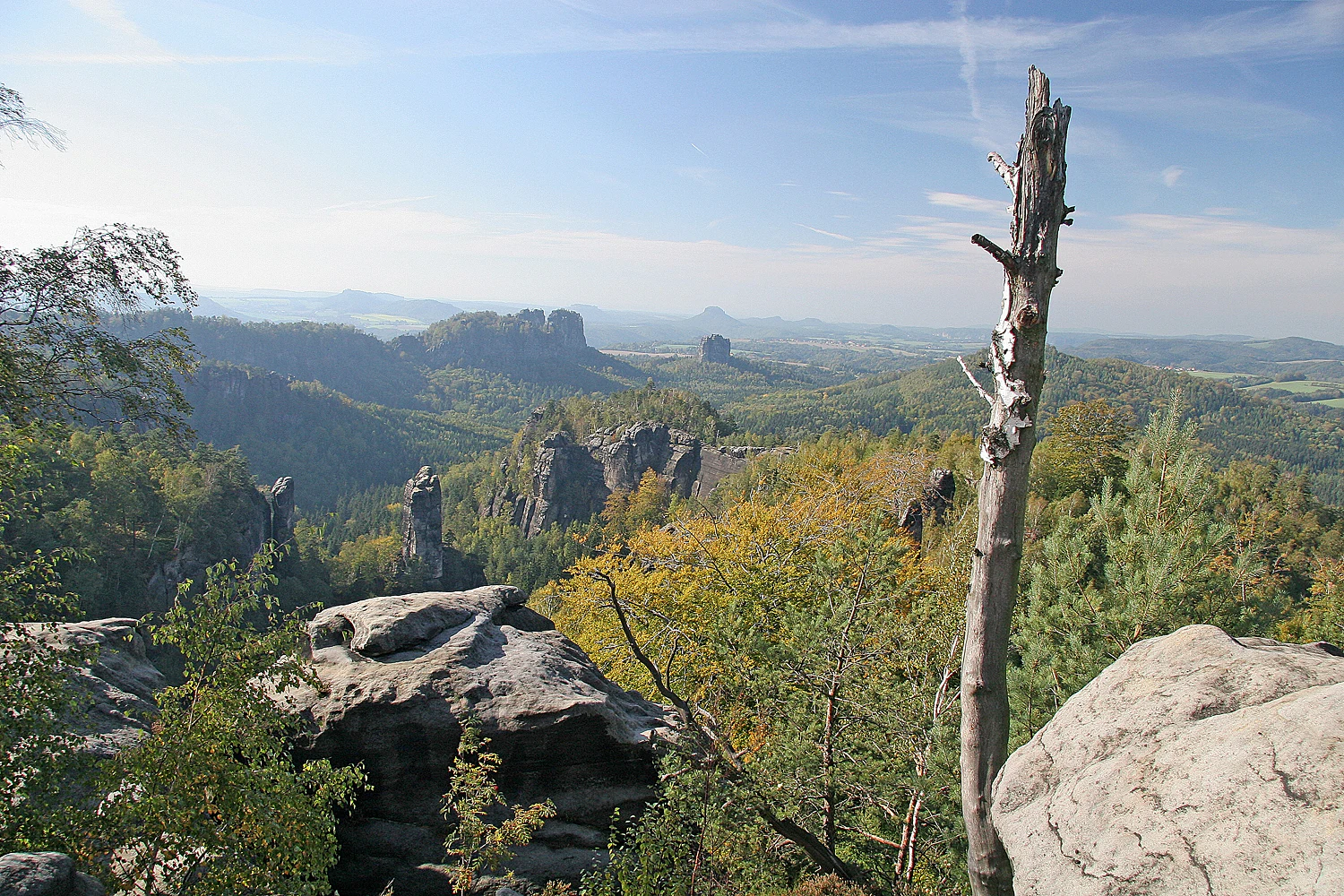 Panoramic view of sandstone rock formations, forested hills, and distant mountains under a clear blue sky