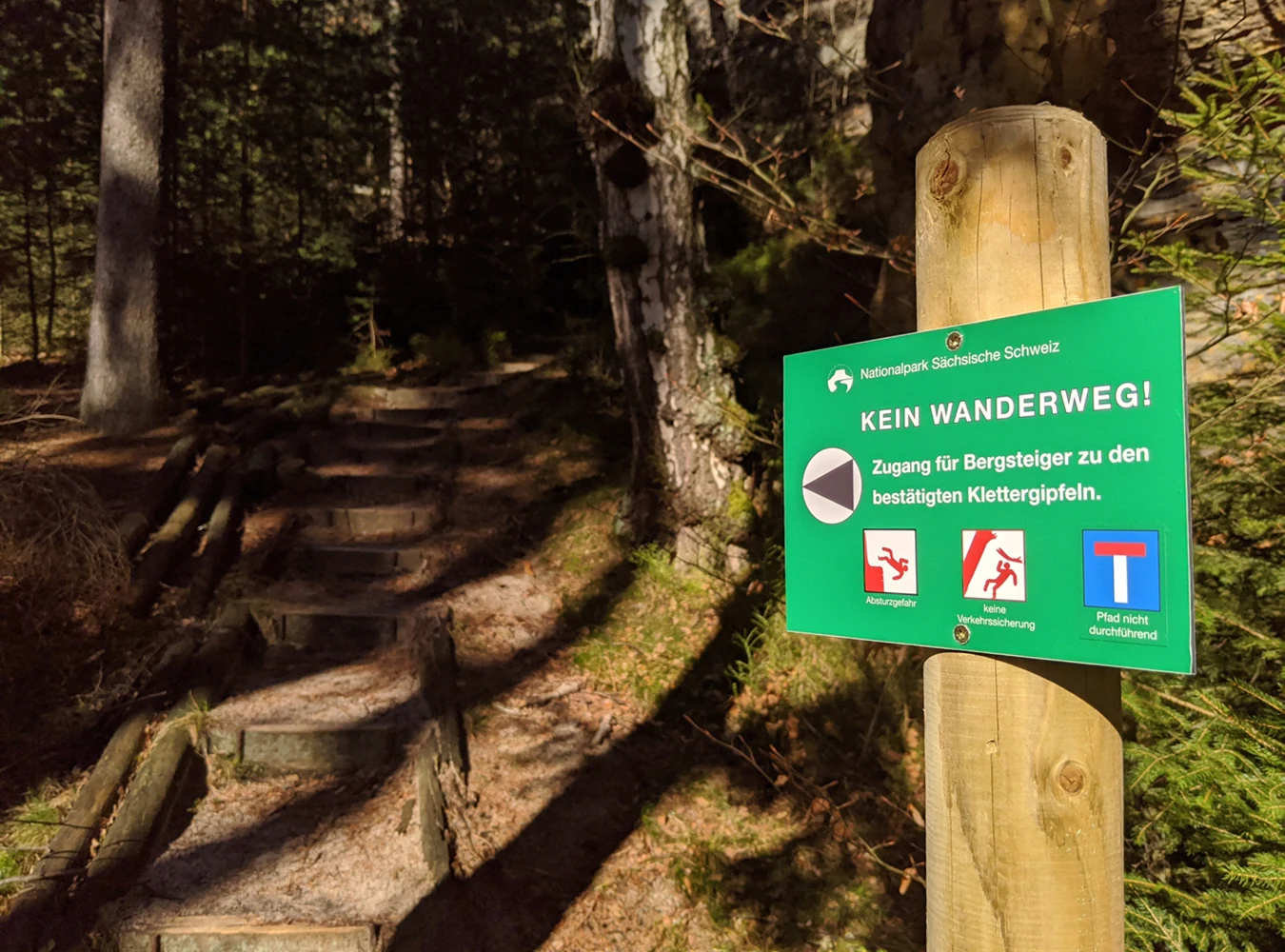 Green sign on wooden post displaying German text 'KEIN WANDERWEG!' with climbing access symbols, set in a forested hiking trail with wooden steps and trees