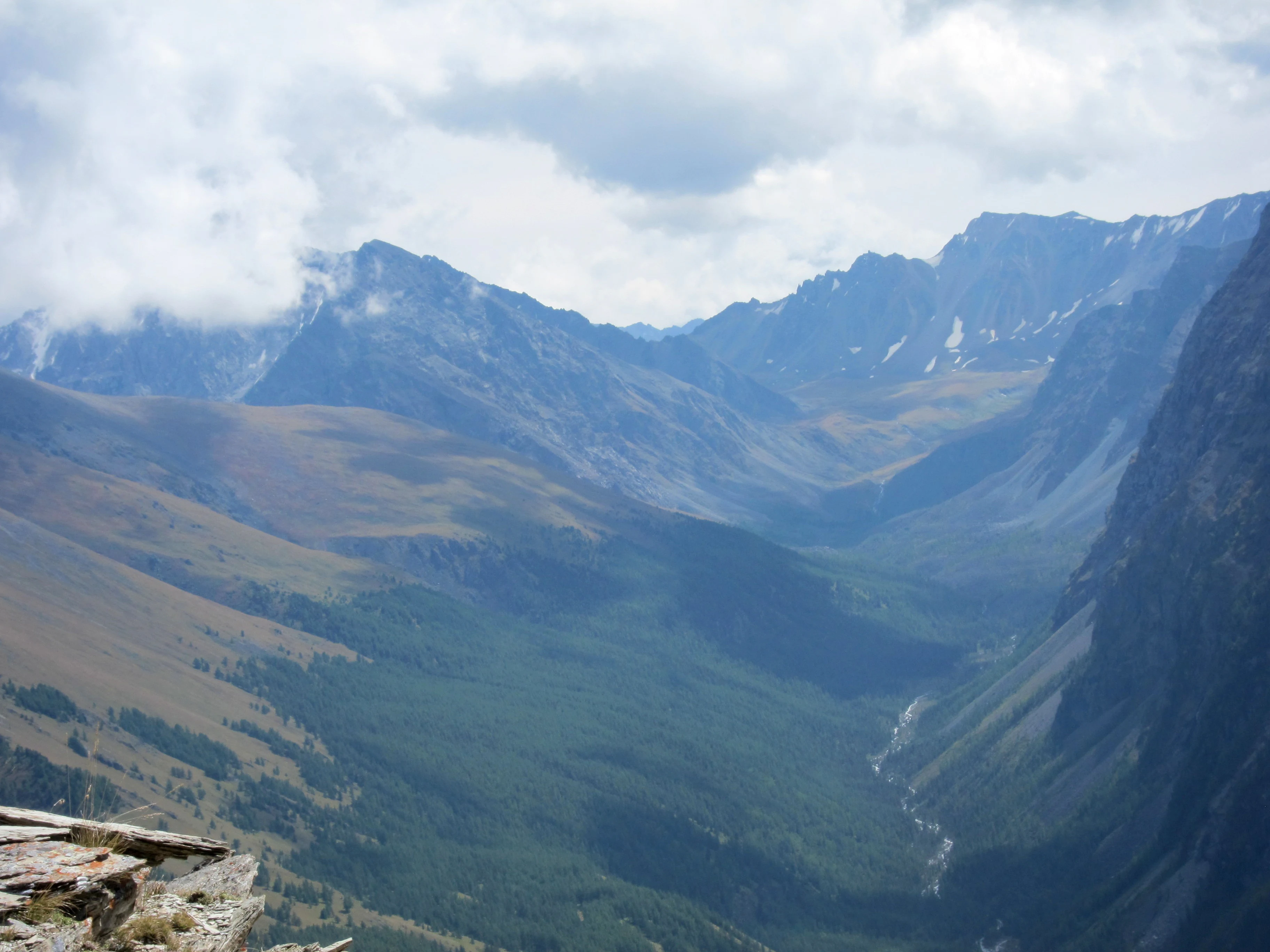 Panoramic view of a mountain valley with river, rocky cliffs, and distant peaks under partly cloudy sky