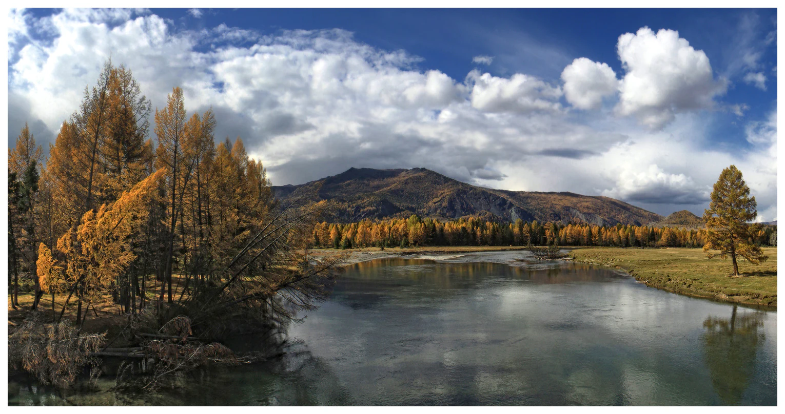 A wide landscape view of a calm river flowing through a valley with autumn-colored trees, mountains in the background, and partly cloudy sky.