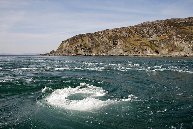 A powerful whirlpool swirling in the deep green waters of the Firth of Lorn, with a rocky, rugged island coastline in the background.