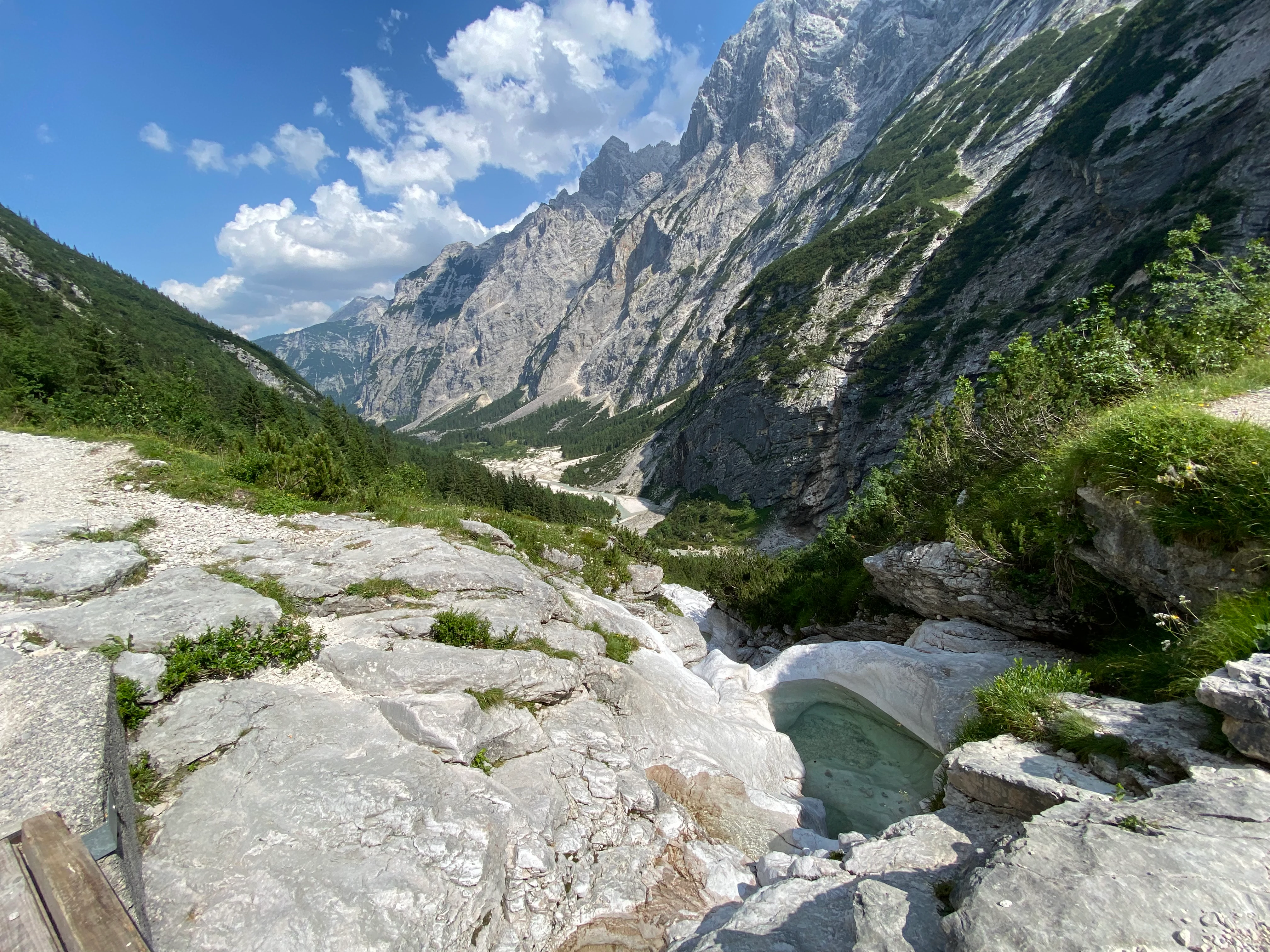 A wide angle landscape shot showing a rocky alpine valley with a small natural pool in the foreground and towering mountain peaks under a blue sky.