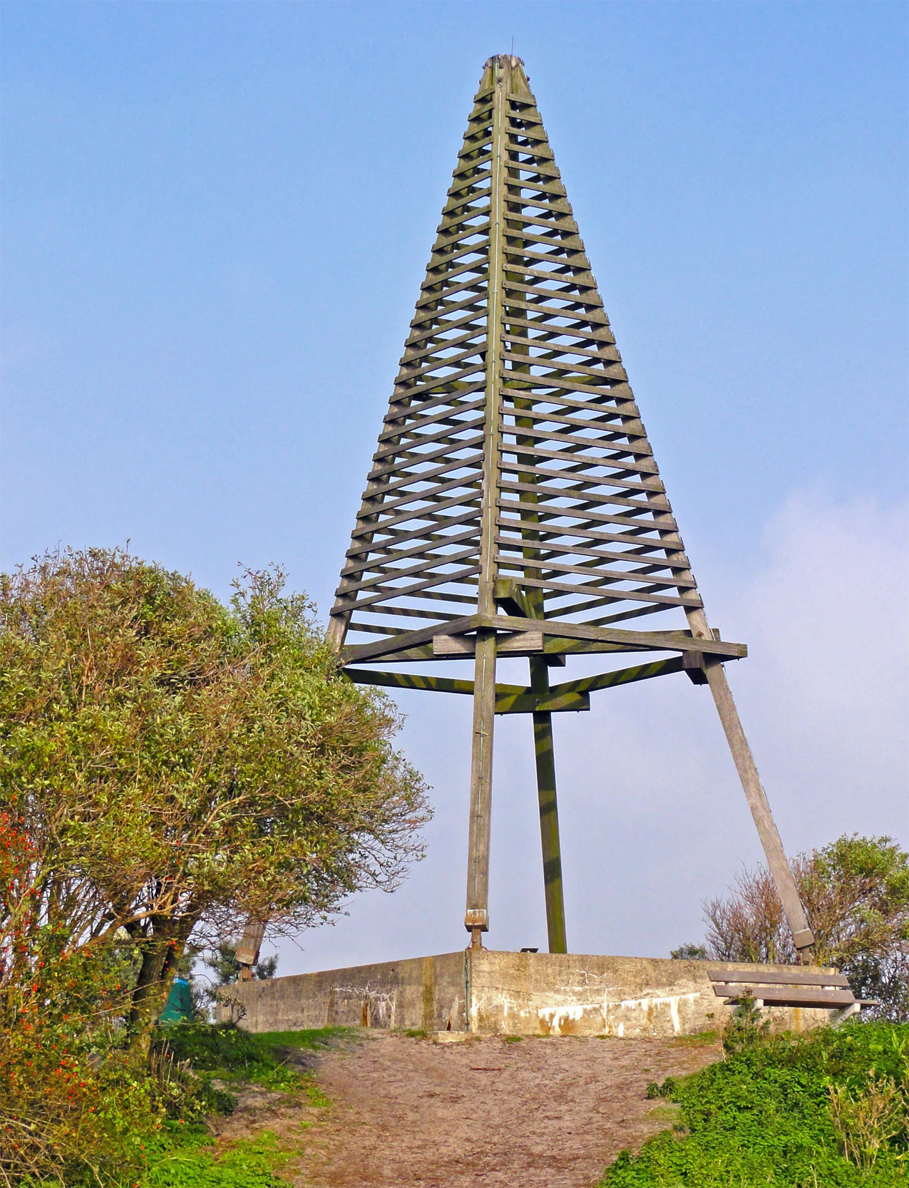 Tall wooden lattice observation tower on a concrete base, surrounded by trees and vegetation under a clear blue sky