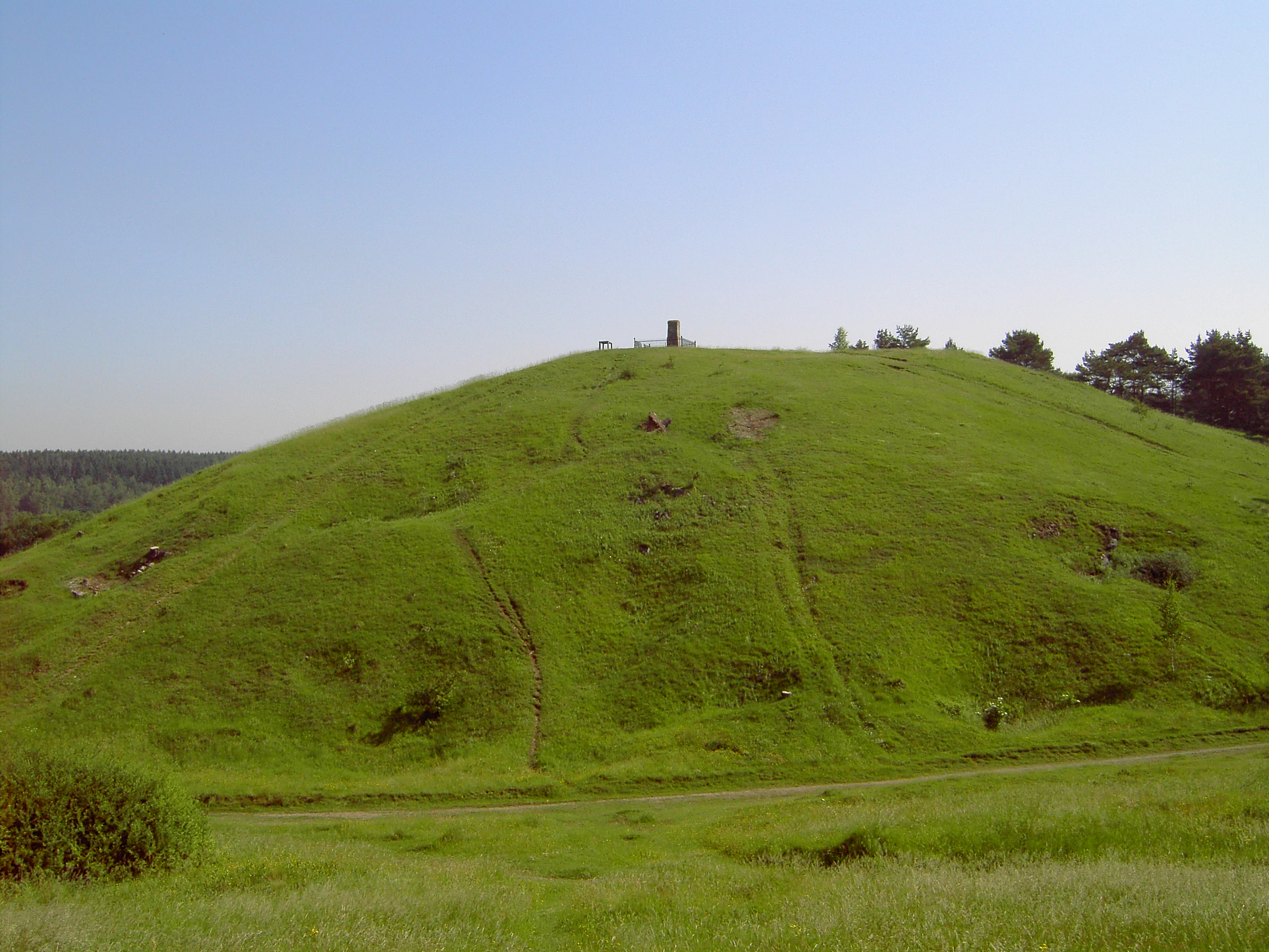 A green, grass-covered hill under a clear sky with a small structure on the summit.