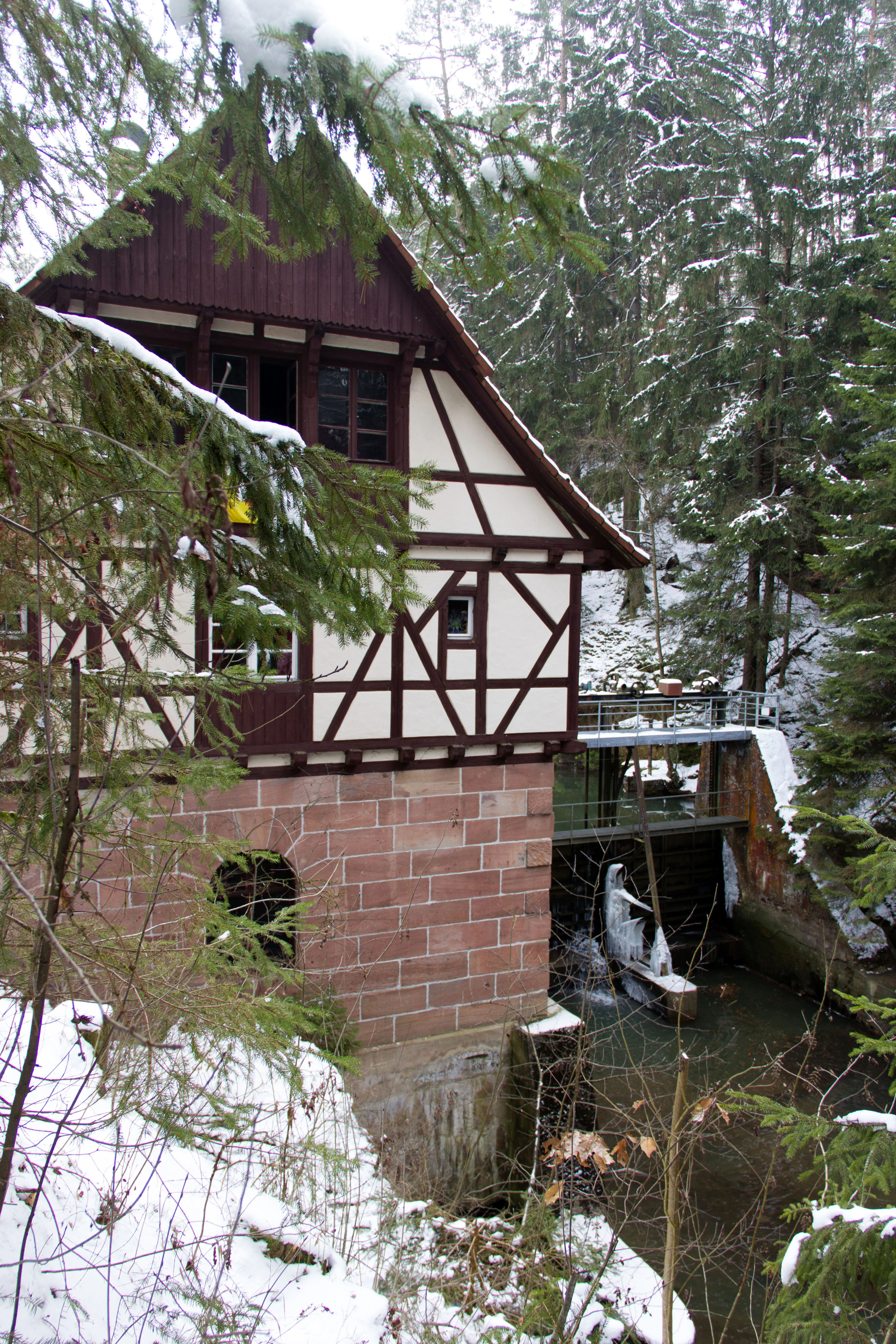 A timber-framed building with a stone base sits next to a river in a snowy, forested gorge.