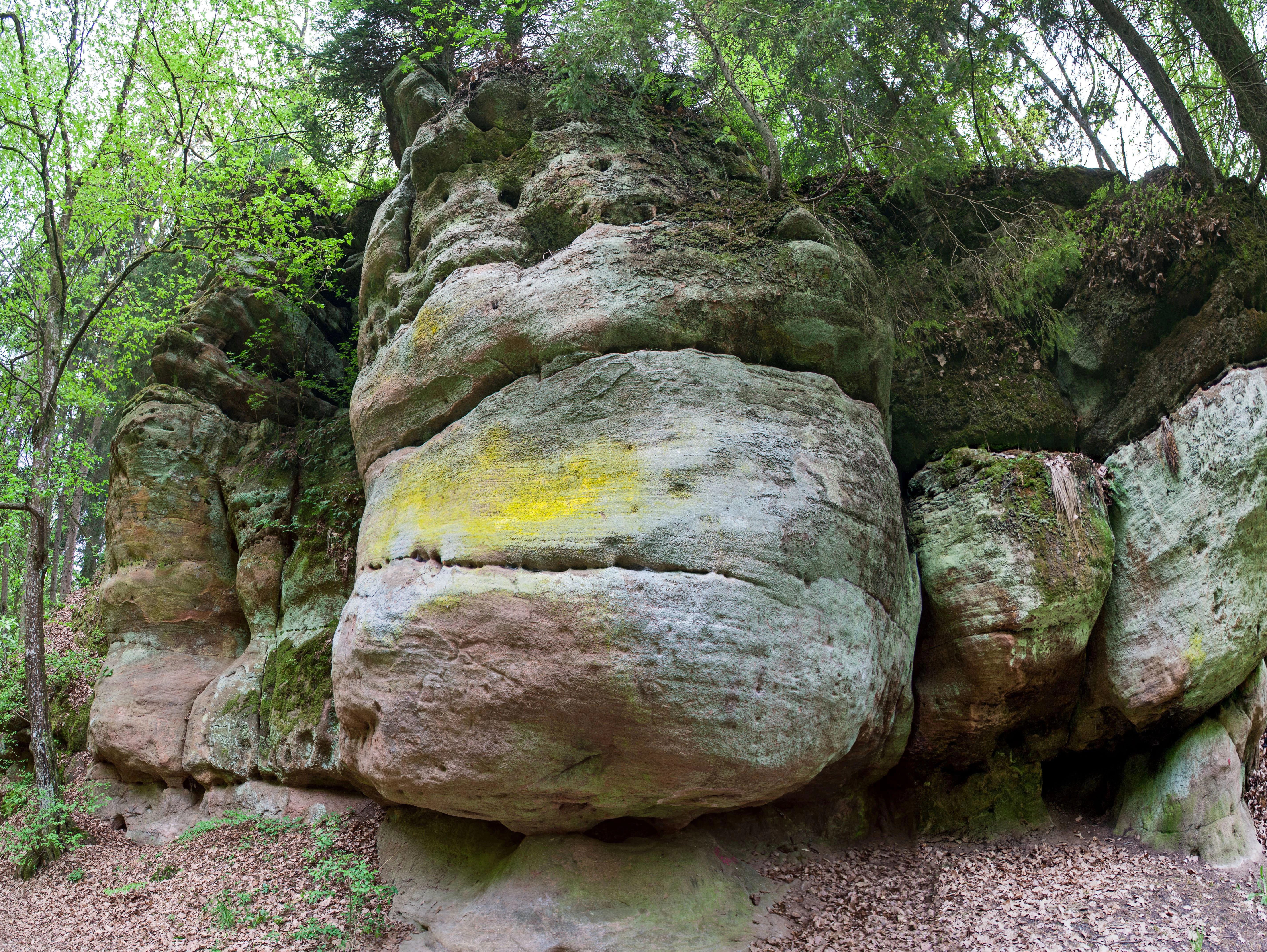 A large, weathered sandstone rock formation covered in moss and lichen, situated within a forest setting.