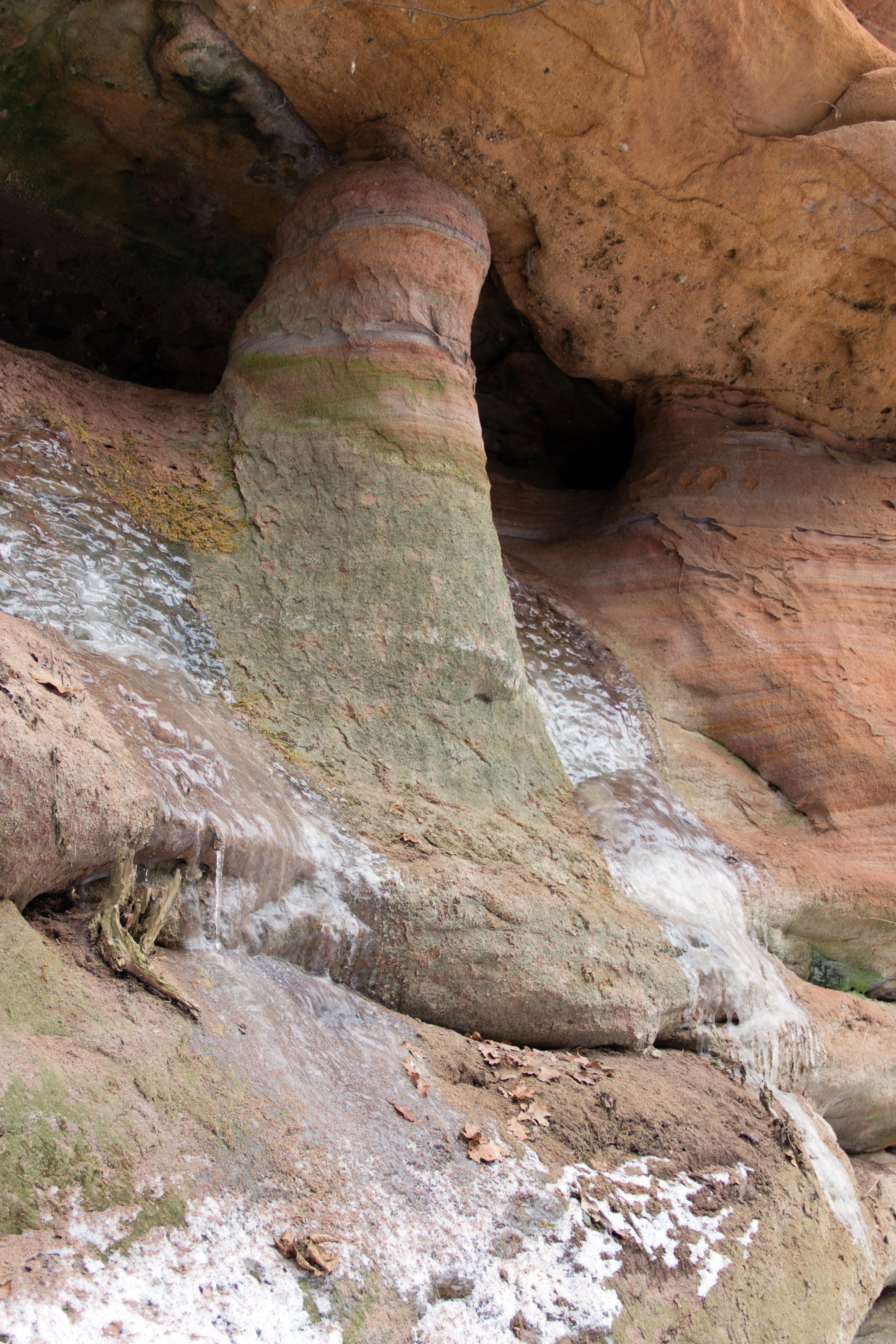 A close-up view of a vertical, cylindrical sandstone rock pillar formation in a gorge with visible mineral streaking and traces of ice or water flow along its surface.