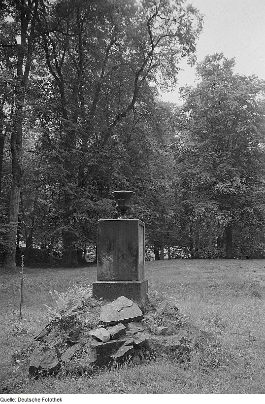 A black and white archival photograph showing a rectangular stone pedestal topped with a small vase, resting on a rocky base in a grassy meadow surrounded by dense deciduous trees.