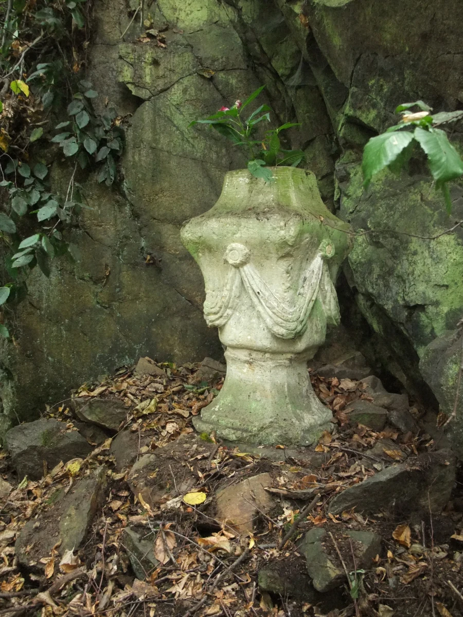 A weathered stone urn monument decorated with carved swags, situated against a mossy rock face covered with ivy and fallen autumn leaves.
