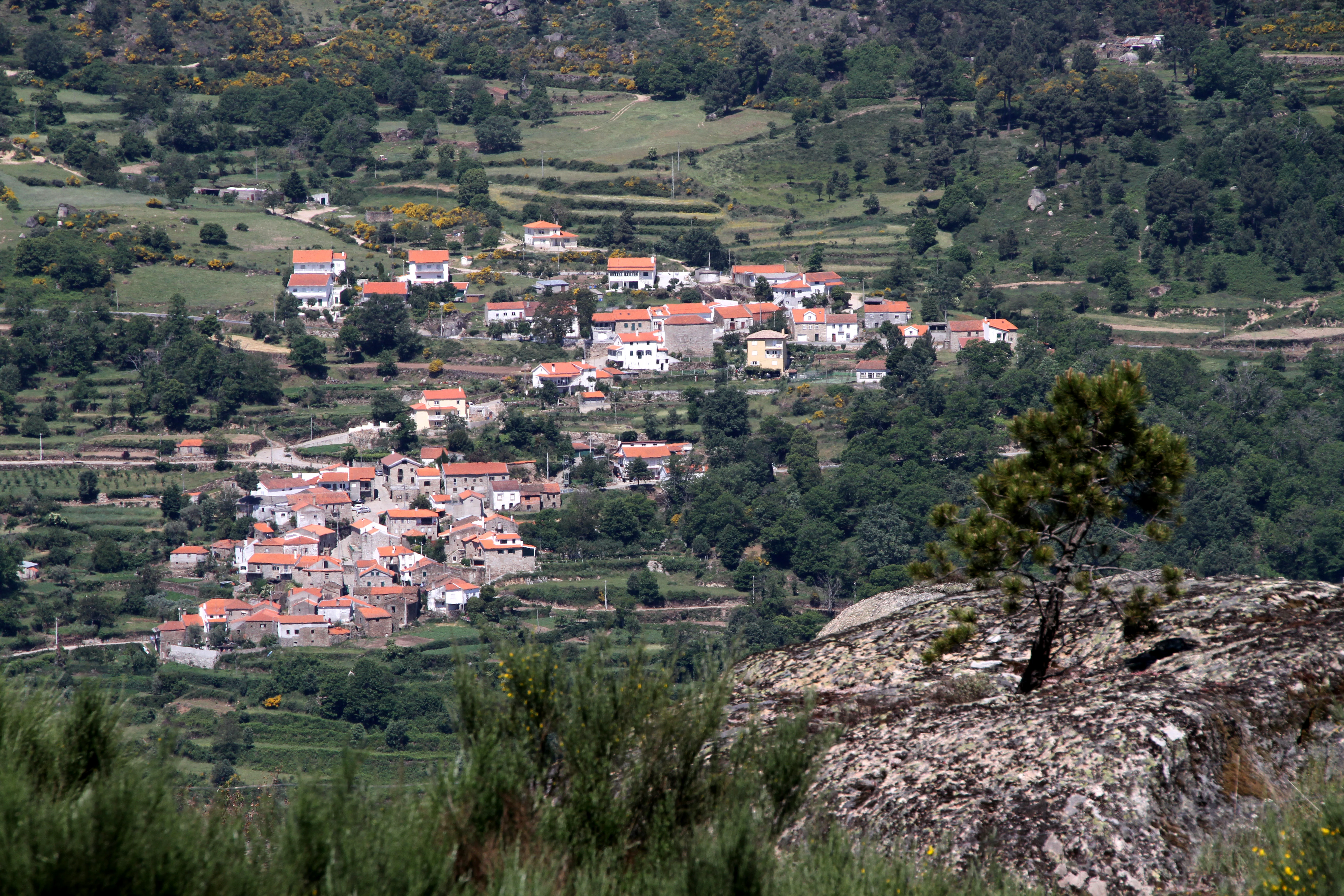 Wide view of a mountain village with white buildings and red roofs surrounded by green hills and rocky terrain