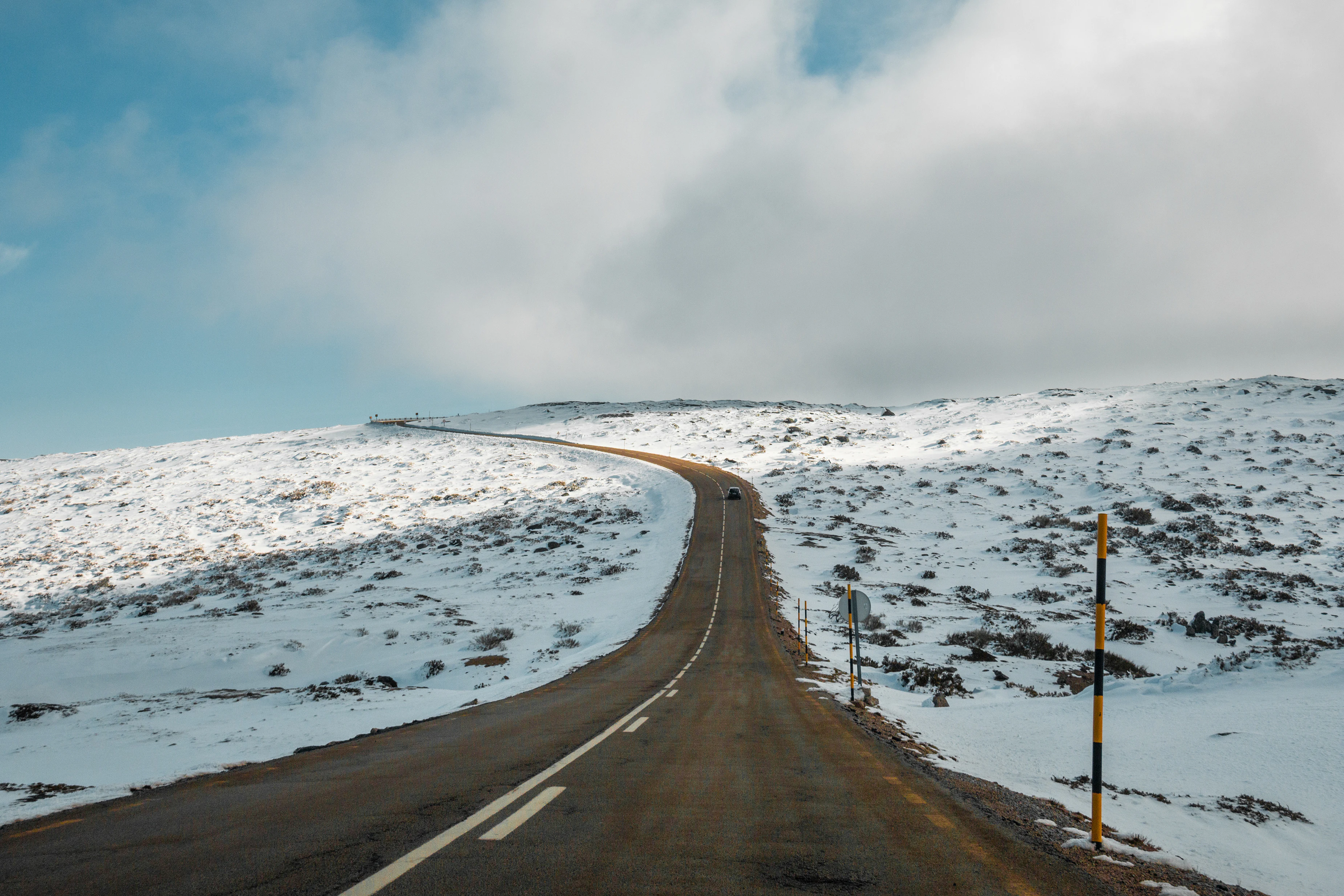 Snow-covered mountain road with yellow and black marker pole, winding through snow-covered hills under partly cloudy sky