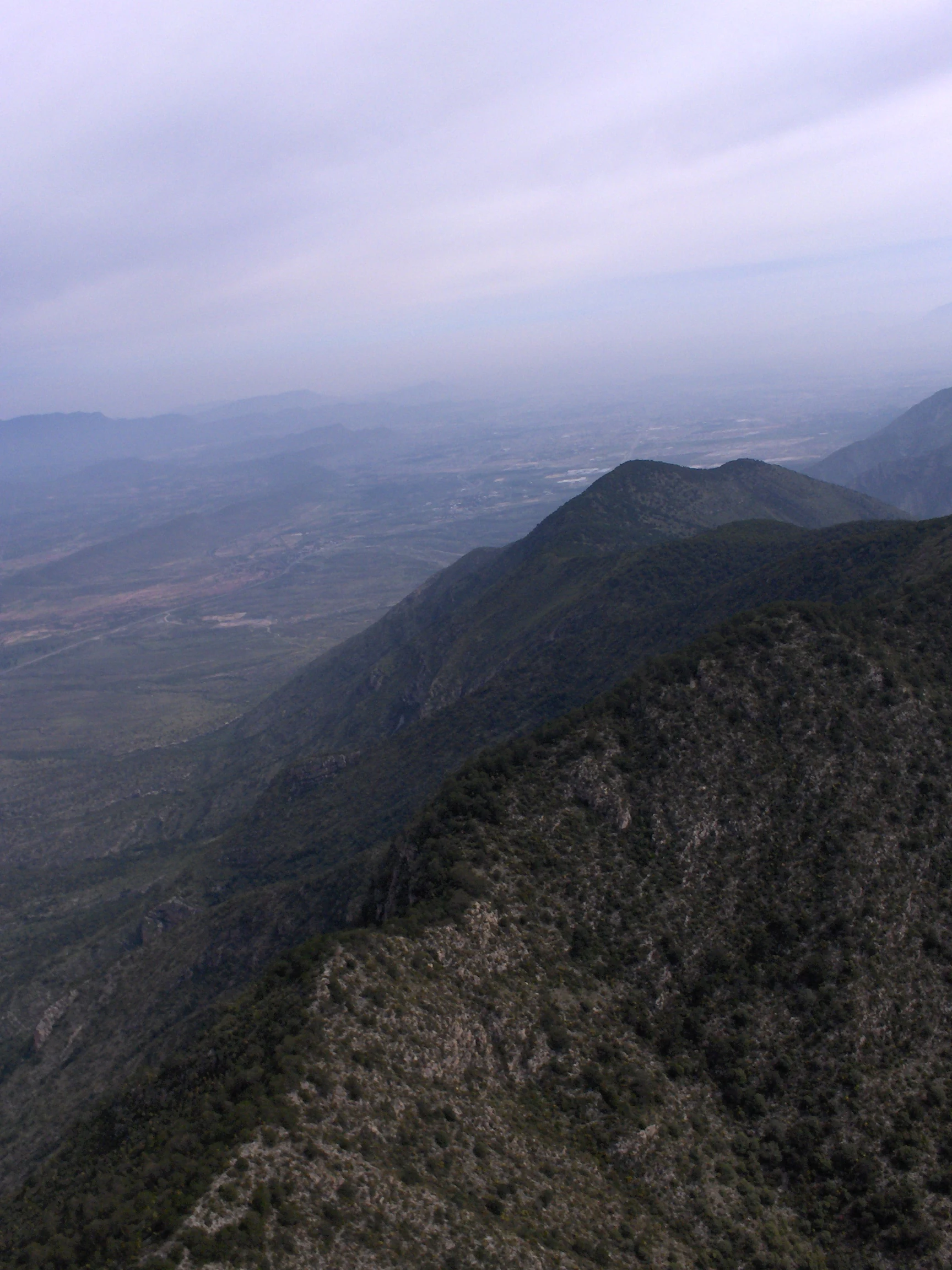 A high-angle view looking down a mountain ridge toward a hazy valley landscape with cloudy skies.