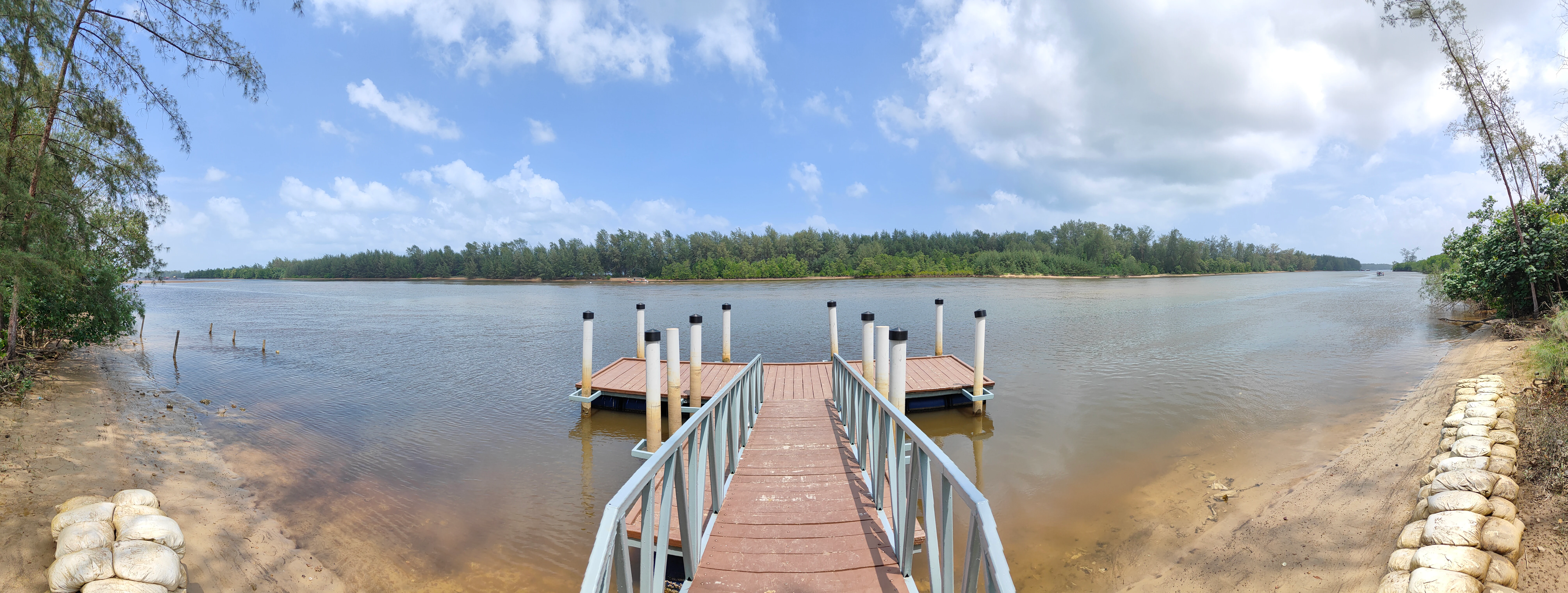 A panoramic photograph of a calm lagoon in Setiu Wetlands State Park, featuring a wooden jetty in the center extending into the water, with a dense forested shoreline in the background.