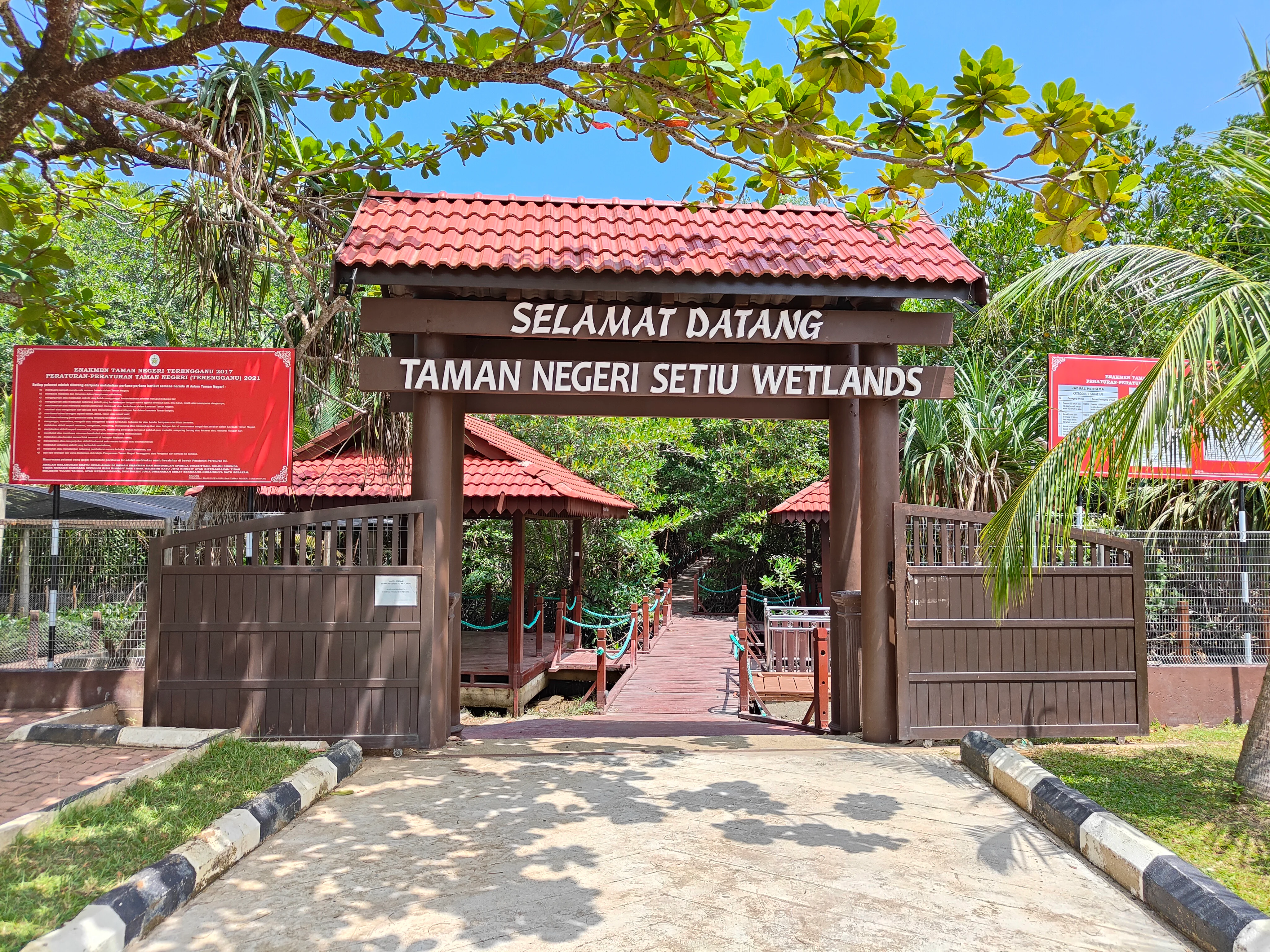 A brown wooden arched gate with a red tiled roof spans a path, featuring signage that reads Selamat Datang Taman Negeri Setiu Wetlands. Red information boards are visible on either side of the entrance gate, set against a backdrop of green trees.