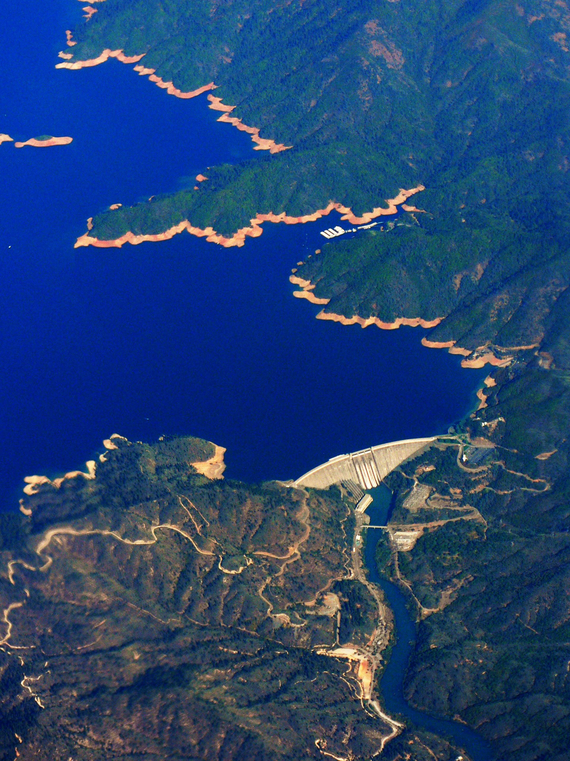Aerial view of the large concrete arch of Shasta Dam holding back the deep blue water of Shasta Lake, surrounded by forested mountains and winding dirt roads.