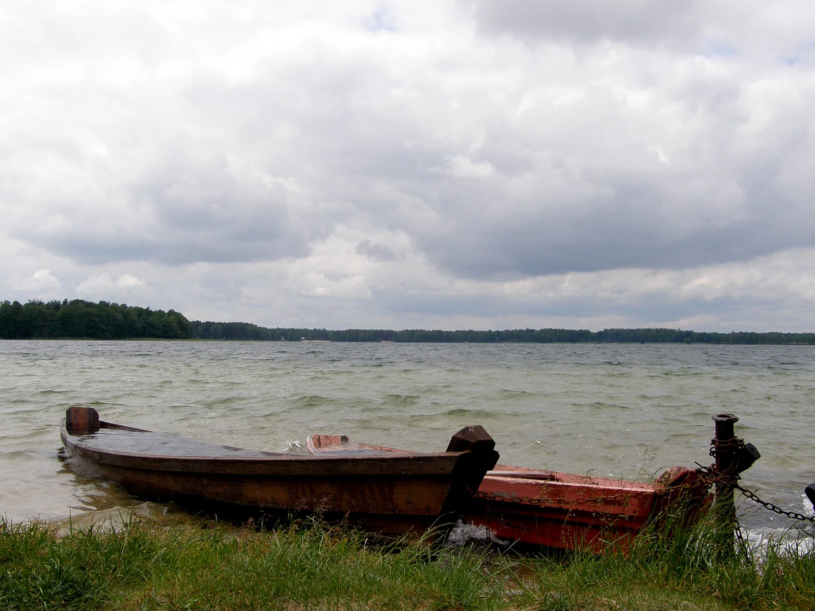 Wooden boat tied to a post on a grassy lake shore under cloudy sky
