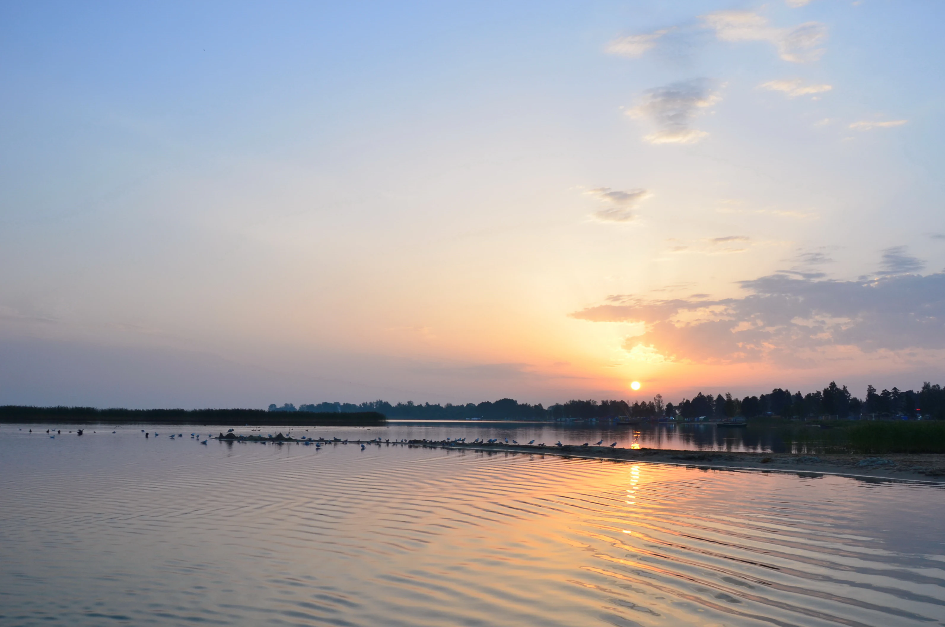 Sunrise over a calm lake with the sun low on the horizon reflecting on the water, distant tree line, and partly cloudy sky