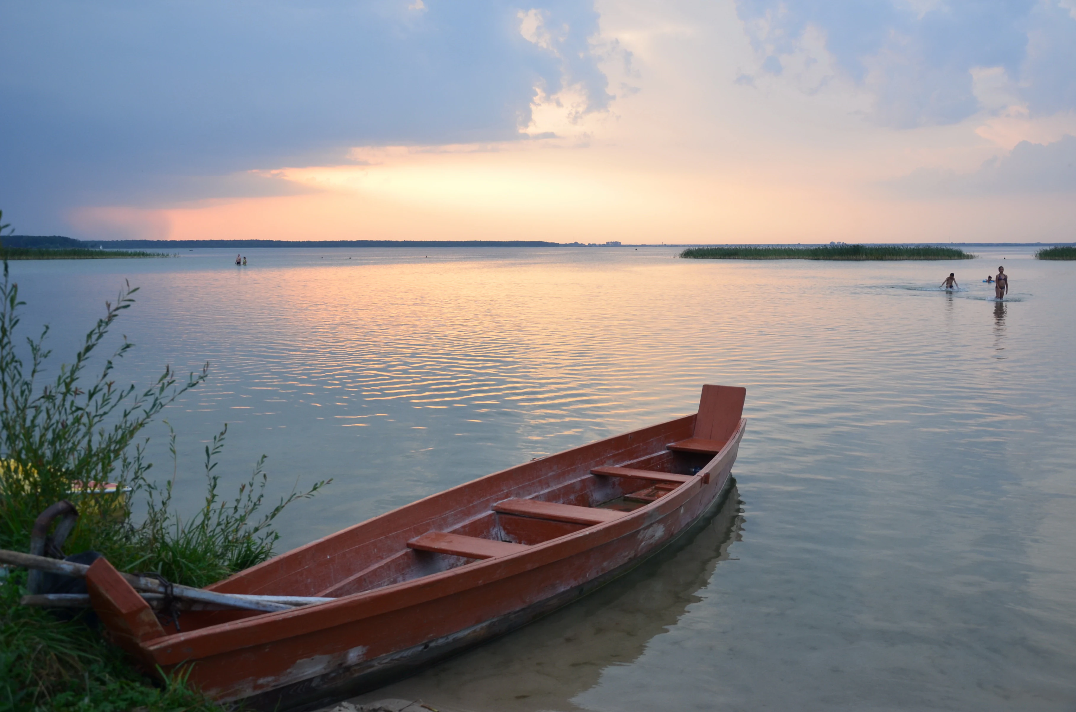 A wooden boat beached on the shore of a lake at sunset with two people wading in the water.