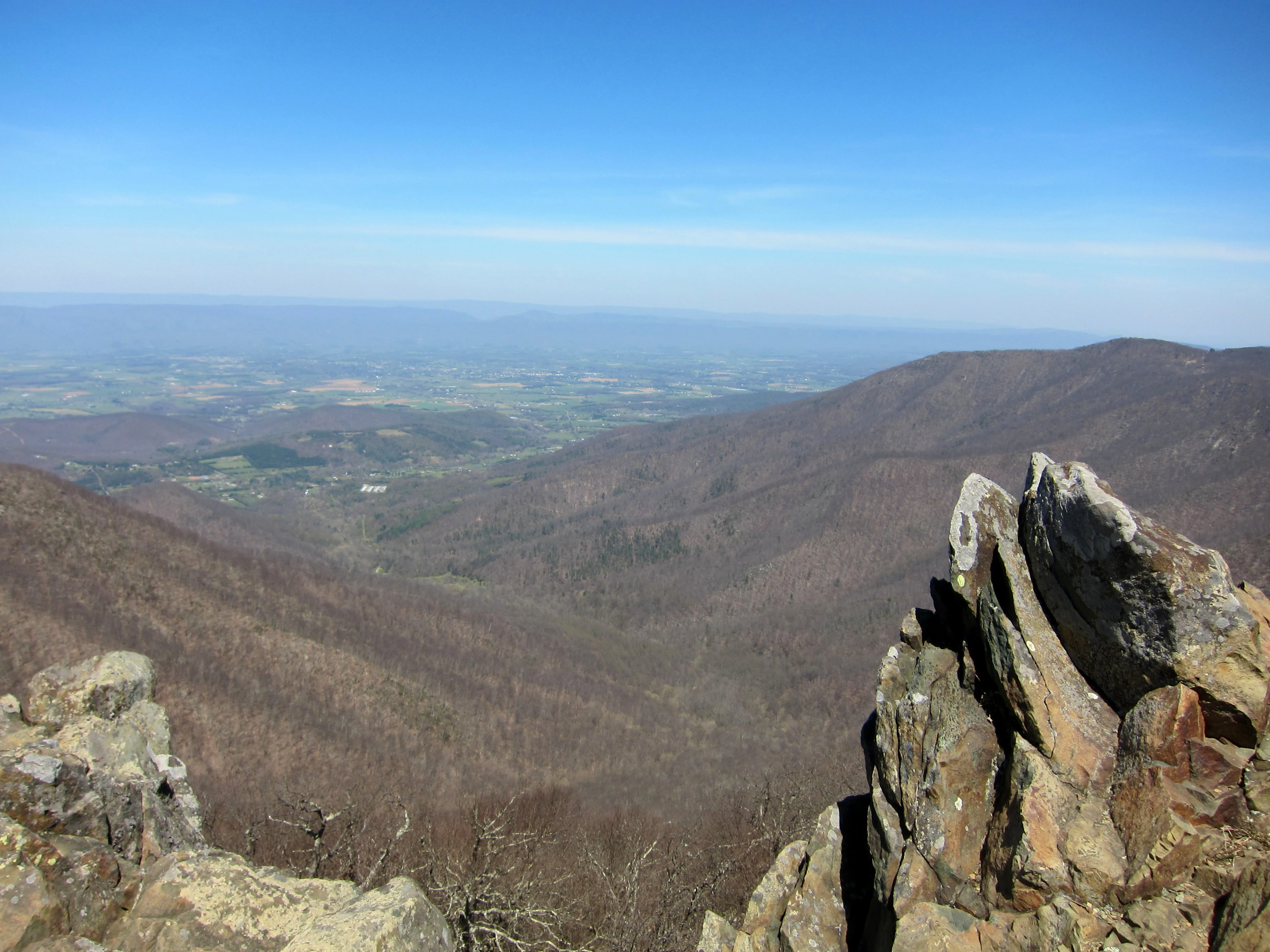 Rocky mountain peak with large boulders overlooking a vast valley and rolling hills under a clear blue sky.