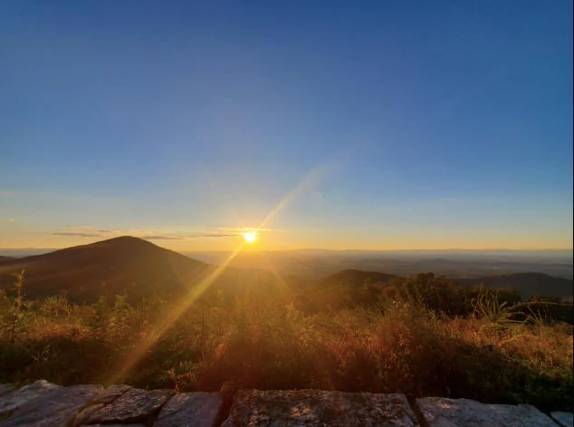 Sunset with golden light illuminating mountains and grassy foreground viewed from a stone overlook